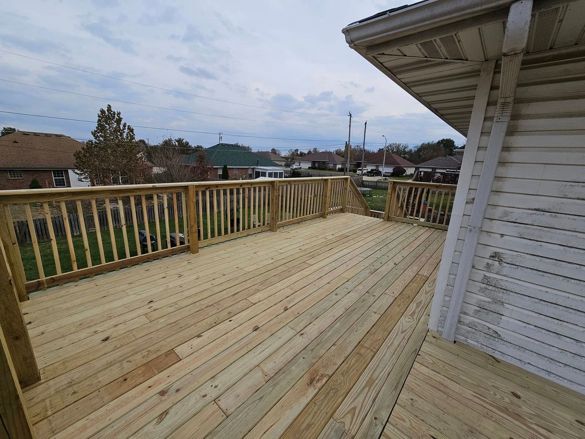 Wooden rooftop deck with railings and a white house wall overlooking a neighborhood at dusk