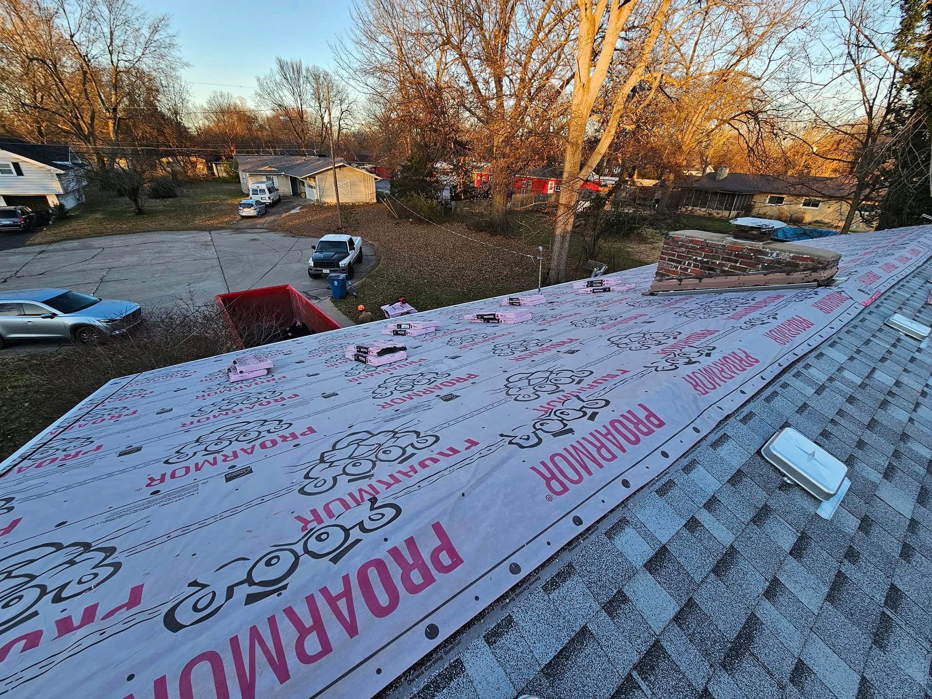 Roof underlayment being installed on a house roof at sunset, with rooftops and trees in the background