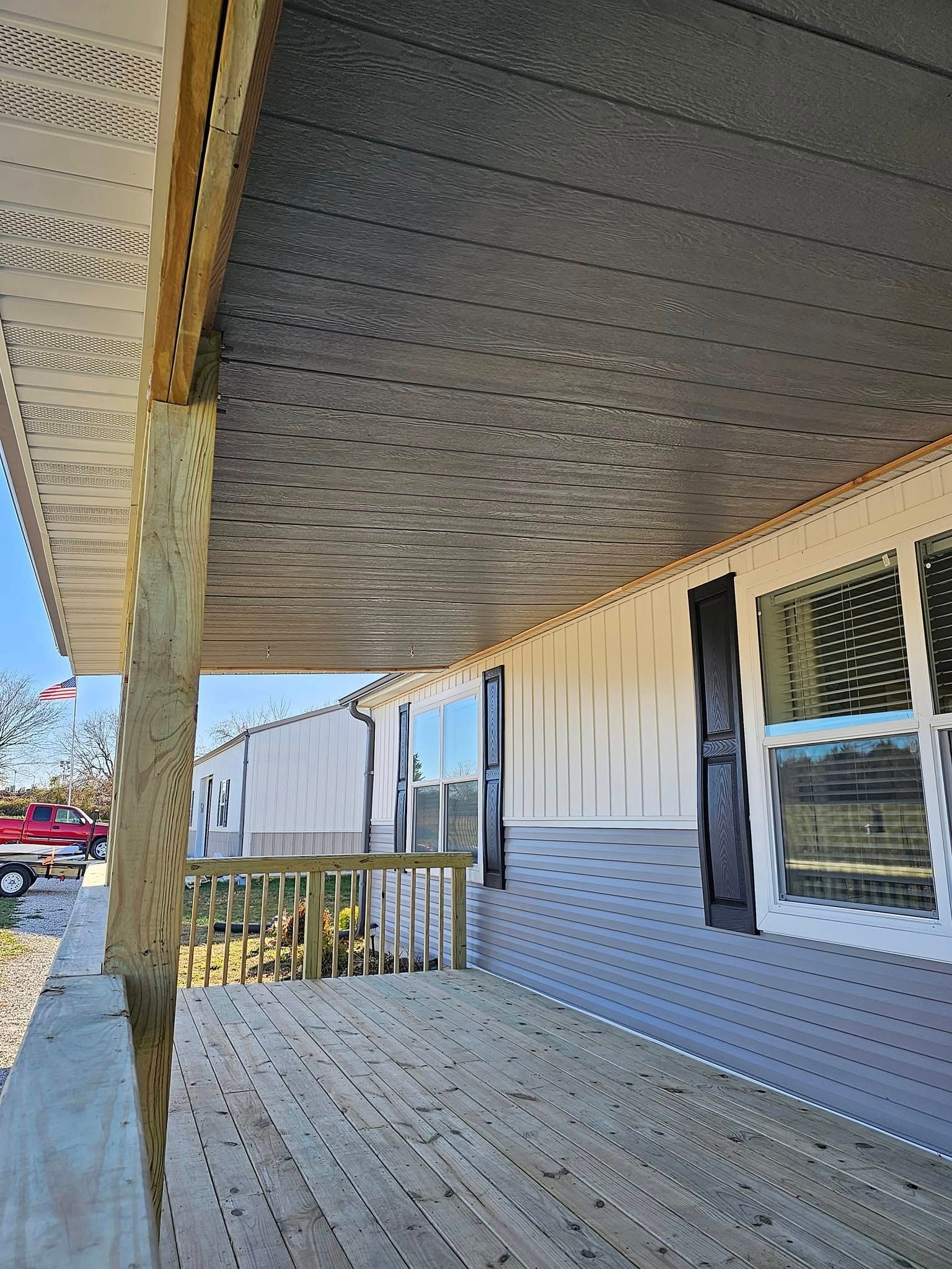 Covered wooden porch with white siding, railing, and window, viewed from a sunlit corner with trees beyond