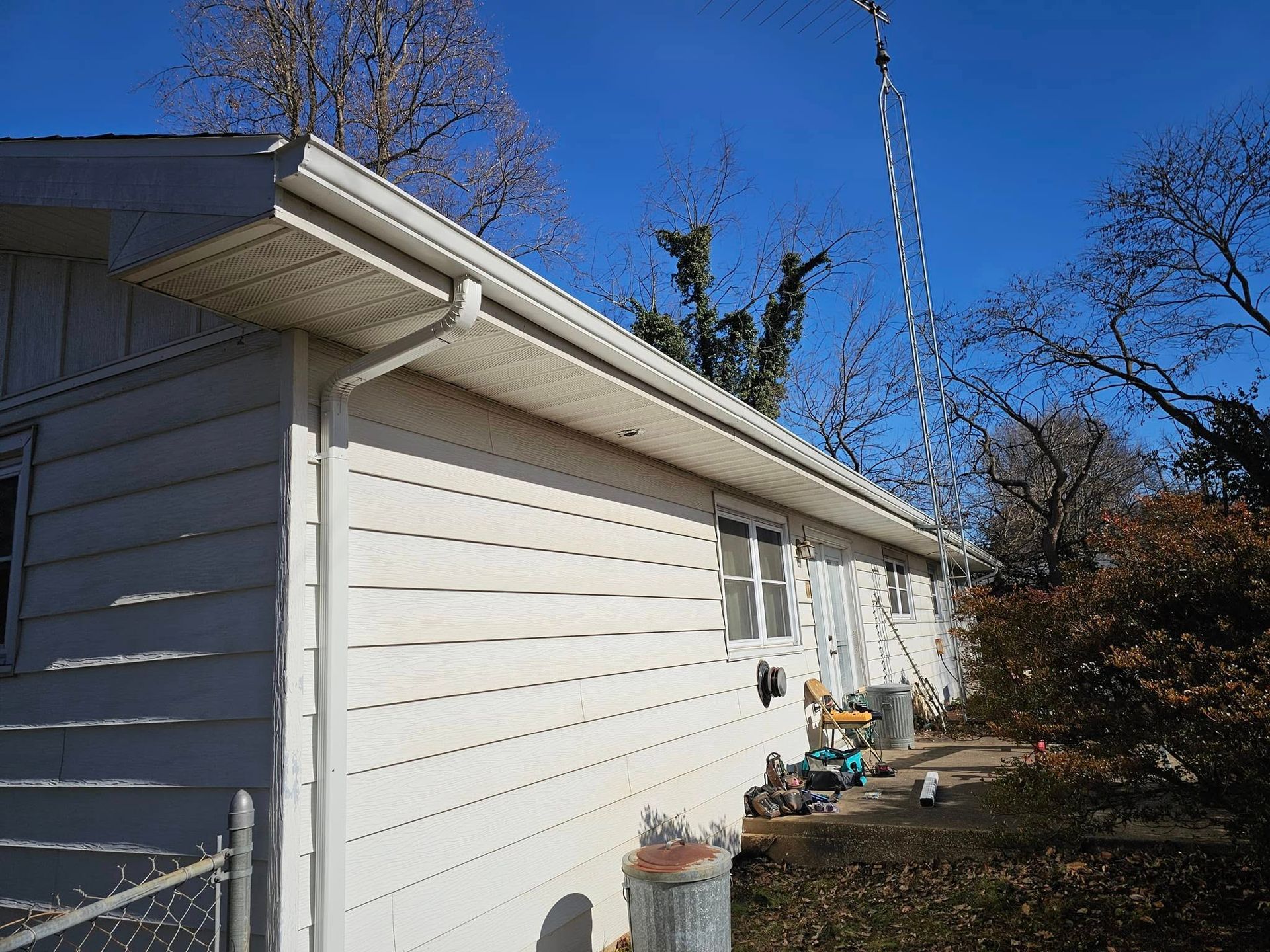 White house siding and roof edge beside a driveway, with trees and yard items under a blue sky