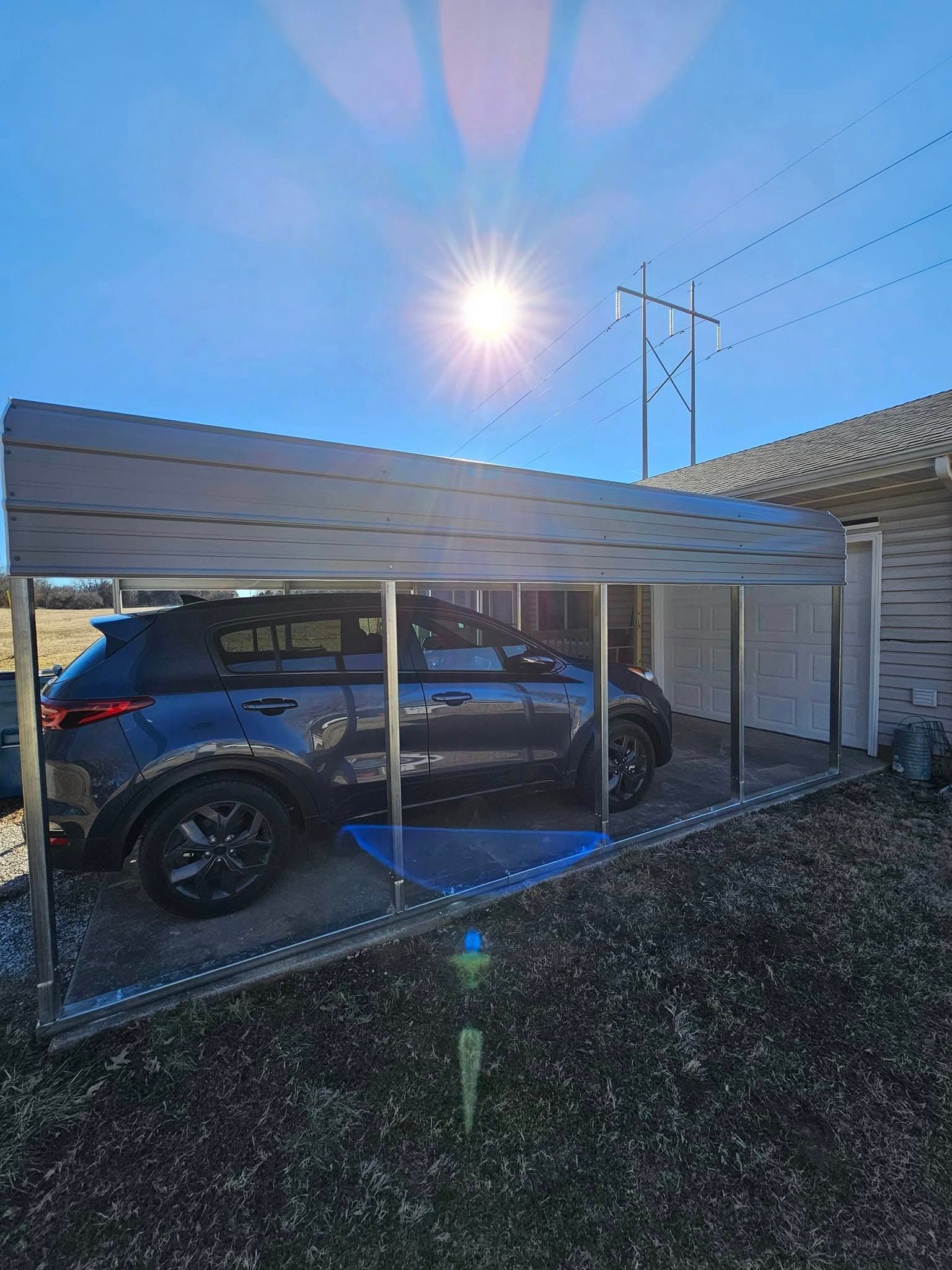Blue SUV parked under a carport in bright sunlight, with chain-link fencing and power lines nearby.
