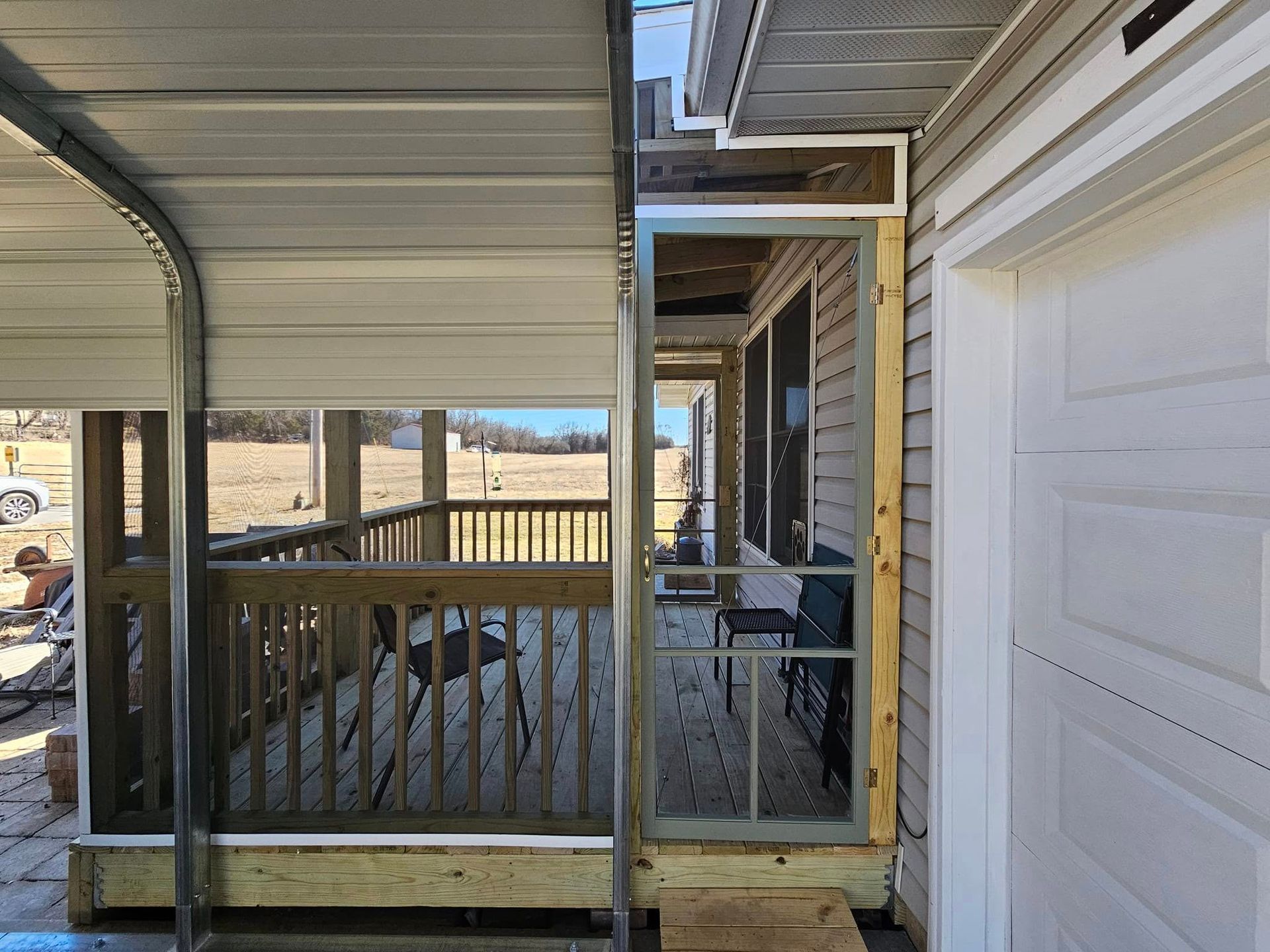 Covered porch with wooden railing, white siding, and a glass door overlooking a yard and open landscape