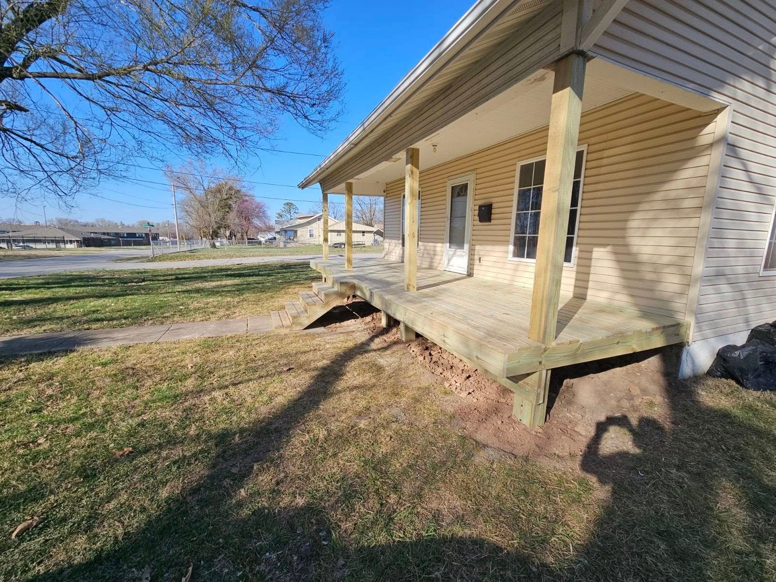 Old beige house with a covered porch and steps, viewed from the yard on a sunny day