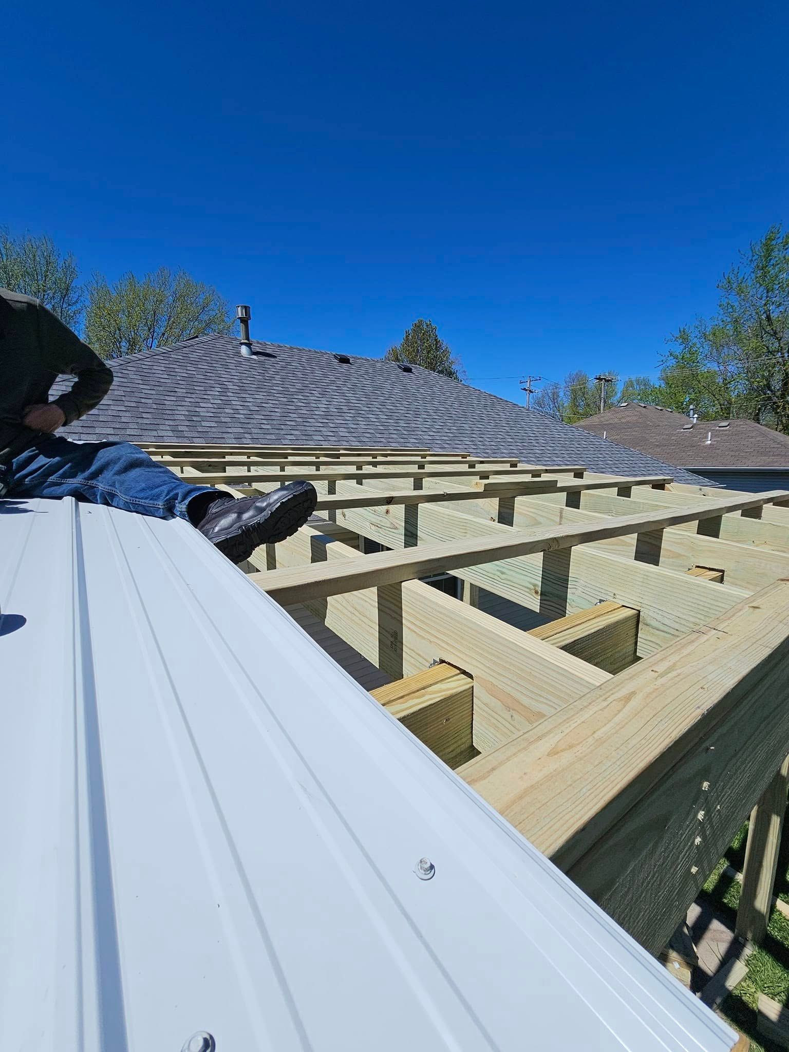 Roof framing under construction beside a gray-tiled roof under a clear blue sky