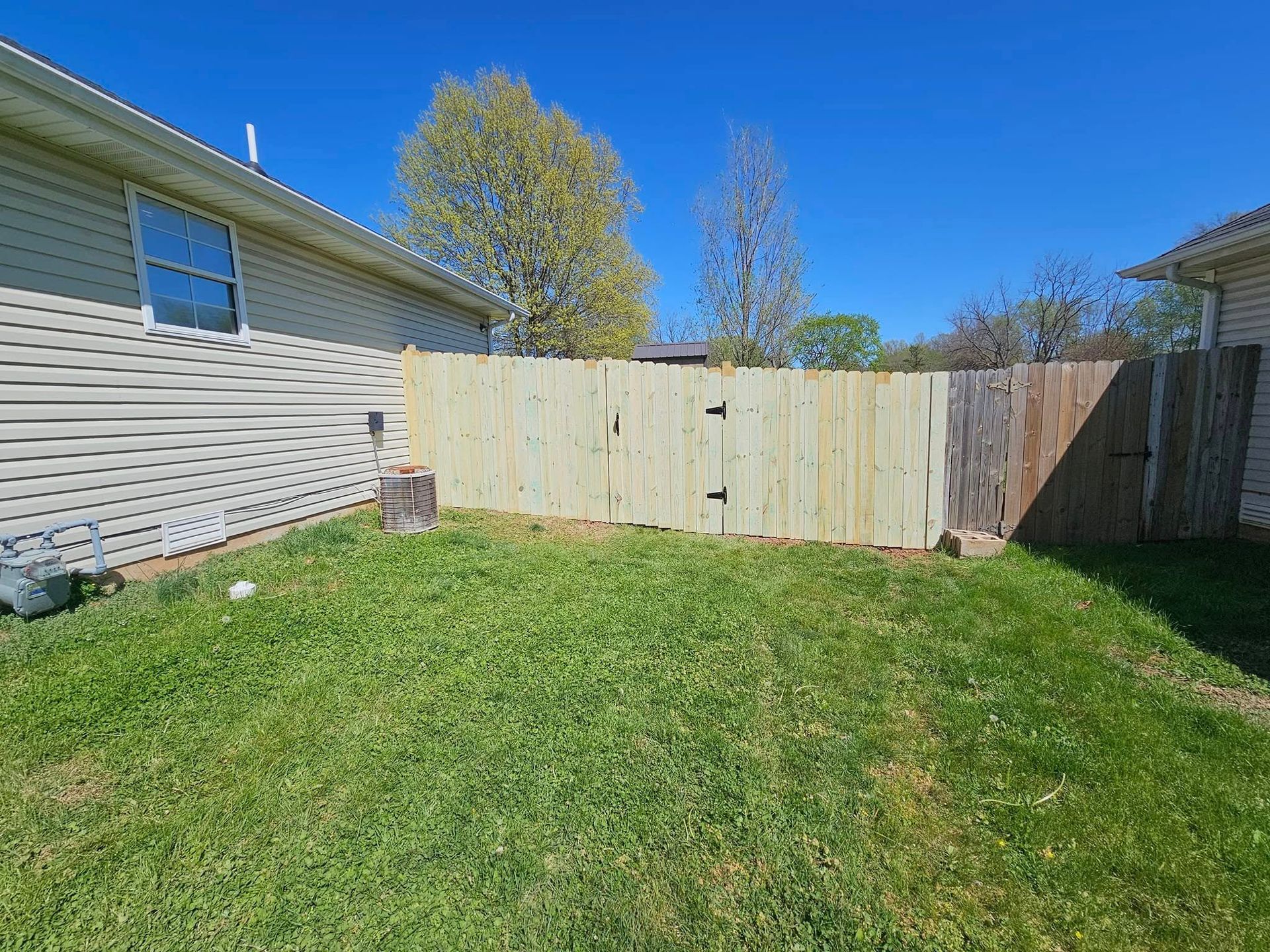 Backyard with green lawn, wooden privacy fence, and house siding under a clear blue sky