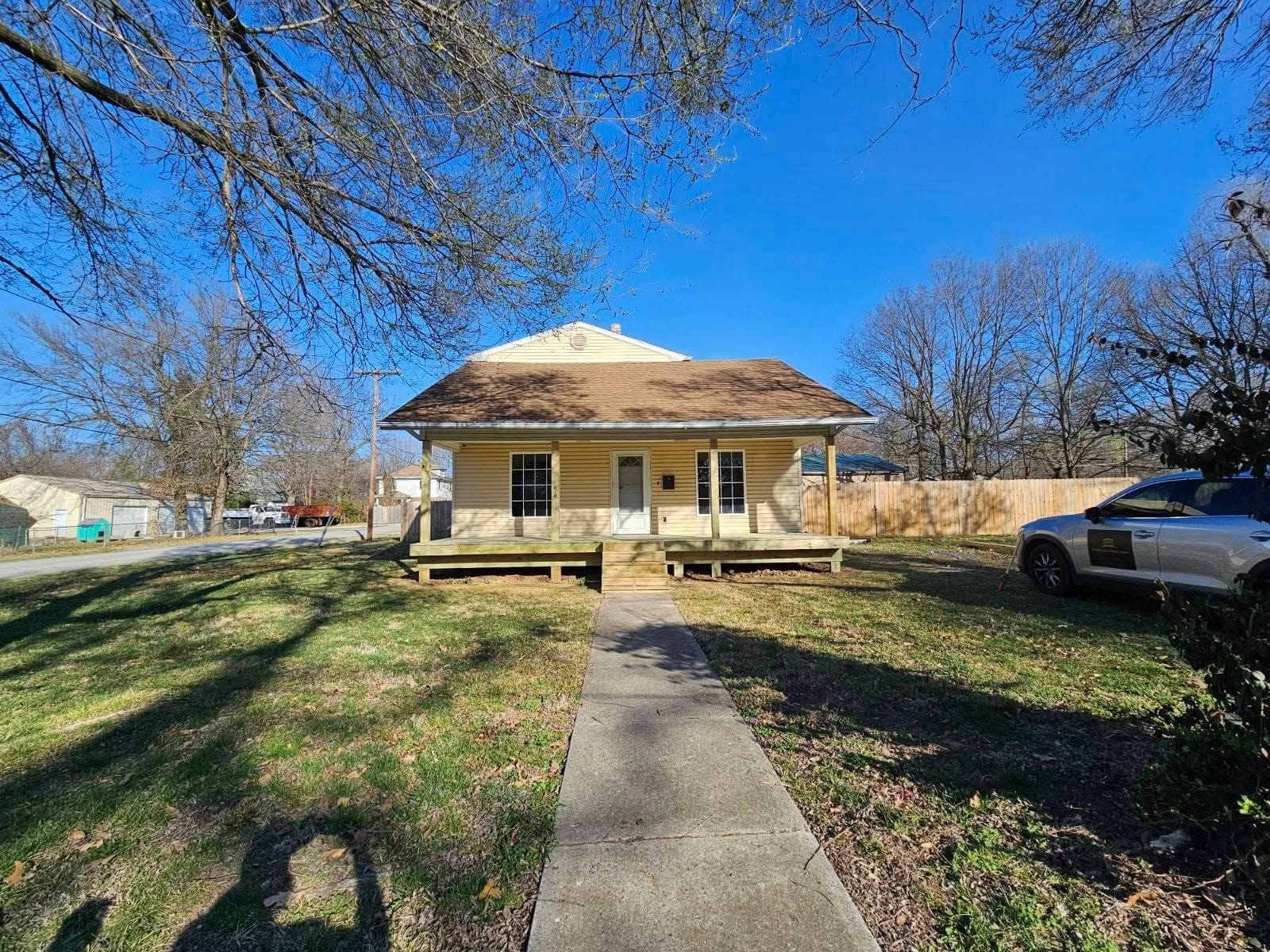 Small beige house with a front porch, centered on a concrete walkway under a clear blue sky