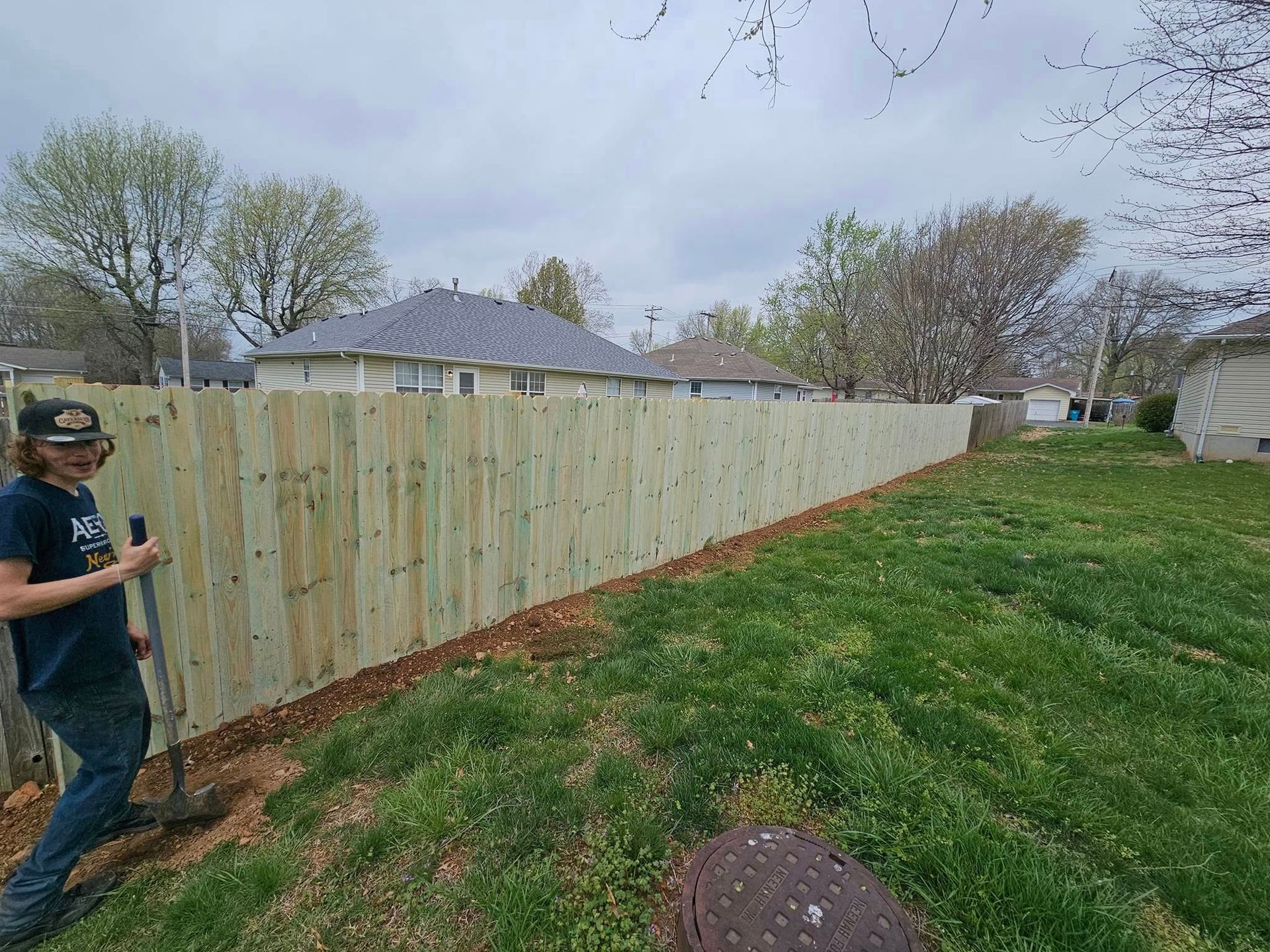 Person standing by a long wooden fence in a grassy suburban yard under cloudy skies