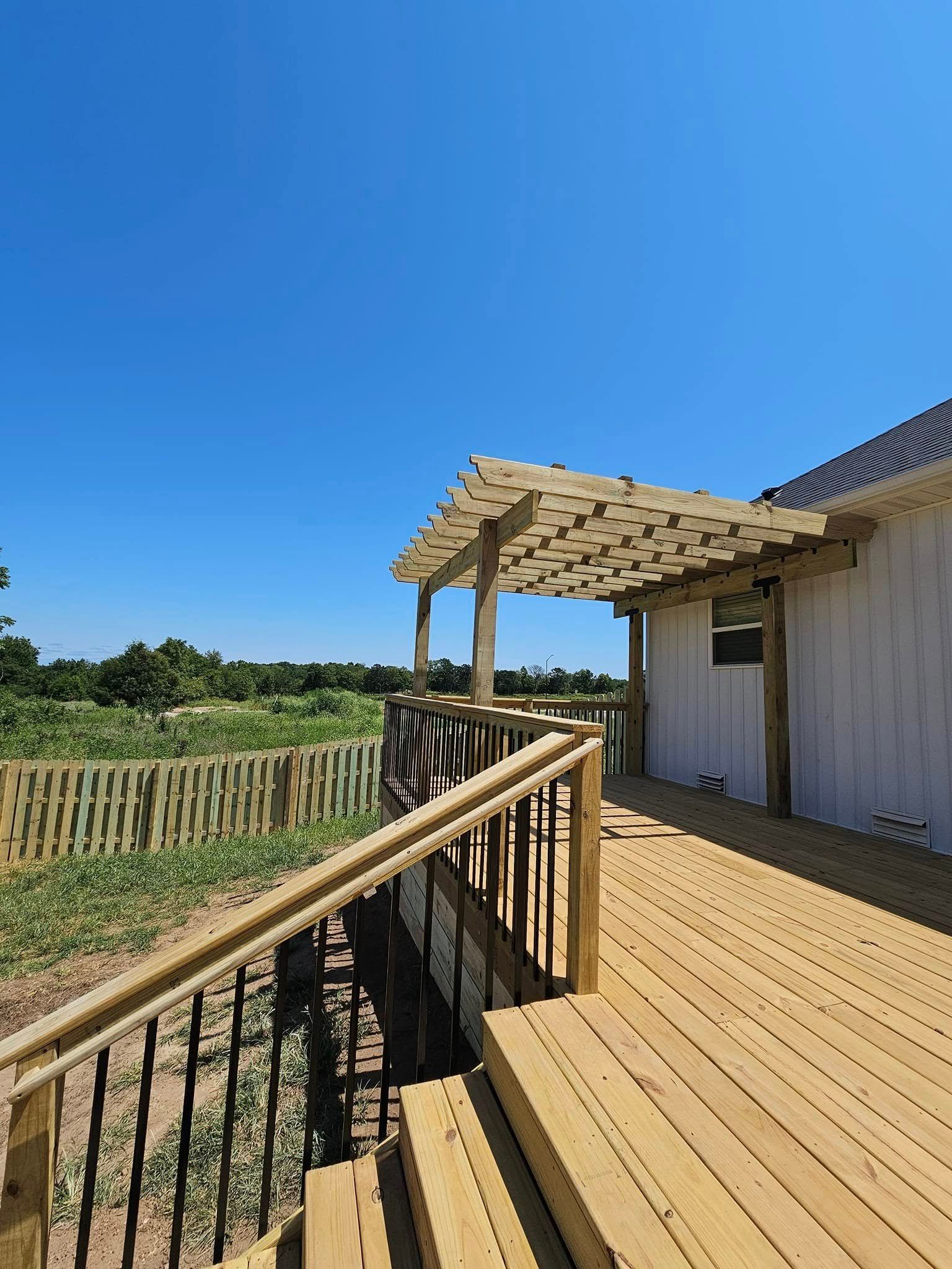 Wooden deck with railing and pergola beside a white building, overlooking a grassy field under a clear blue sky