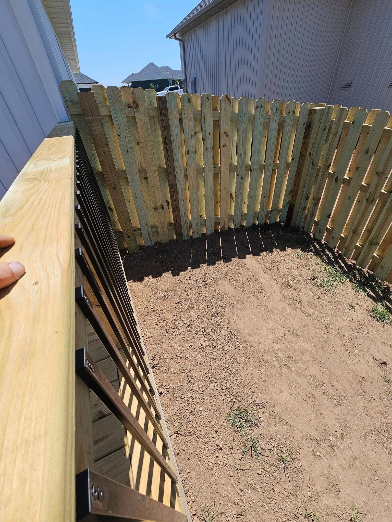 Small fenced dirt yard beside a house, with a wooden gate and slatted privacy fence.
