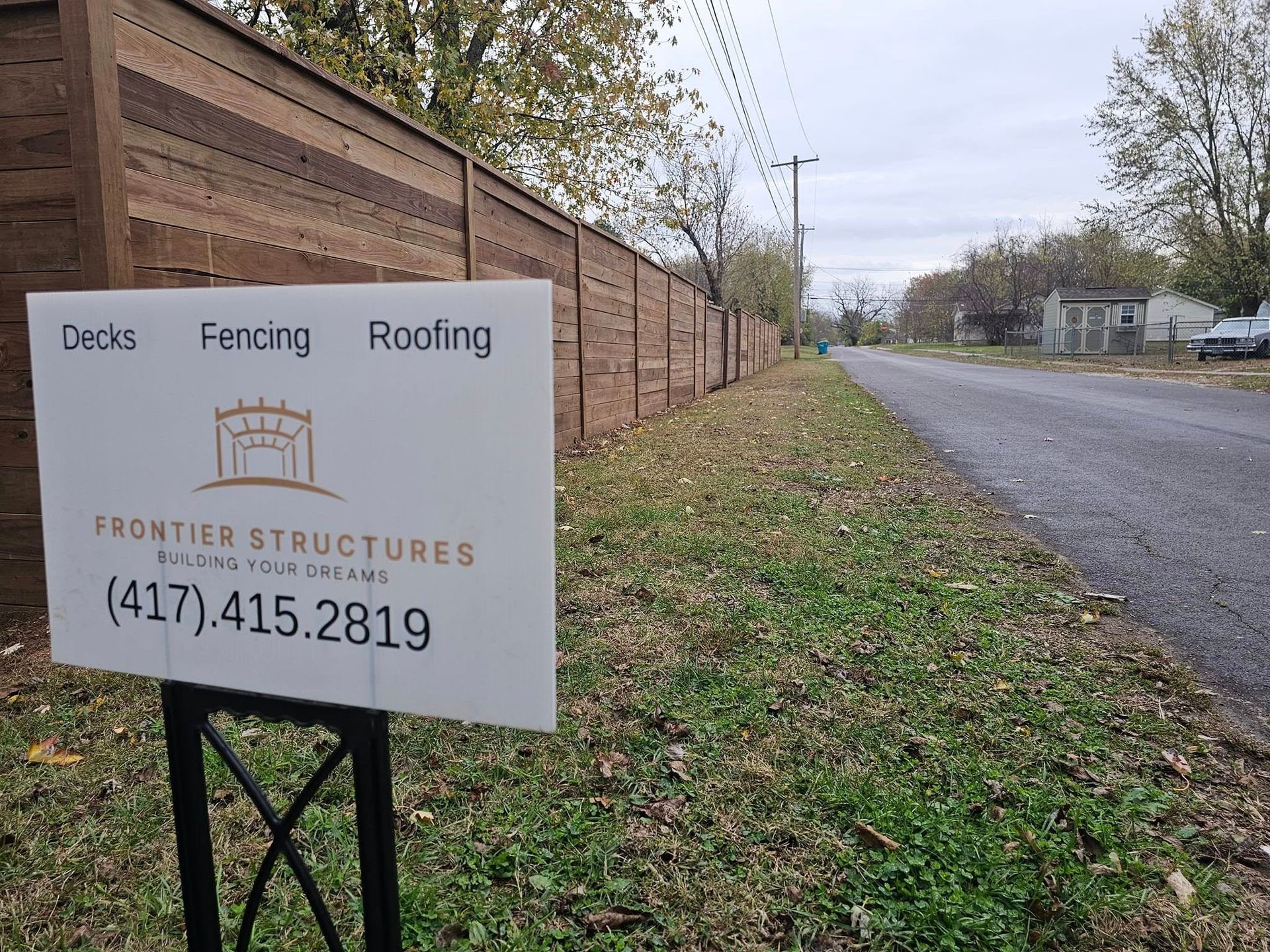 Roadside sign for Frontier Structures beside a brick wall and grassy verge near a quiet street.