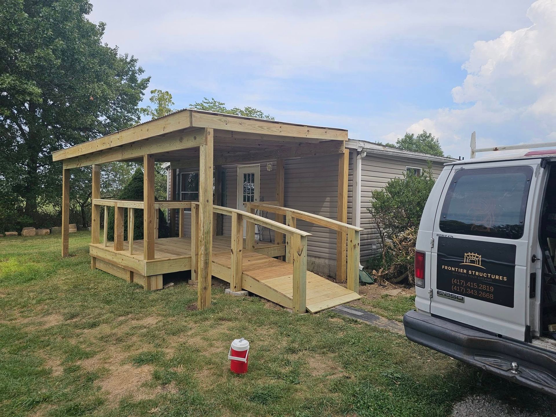Small wooden porch and ramp beside a shed, with a white van parked next to it on a grassy lot.