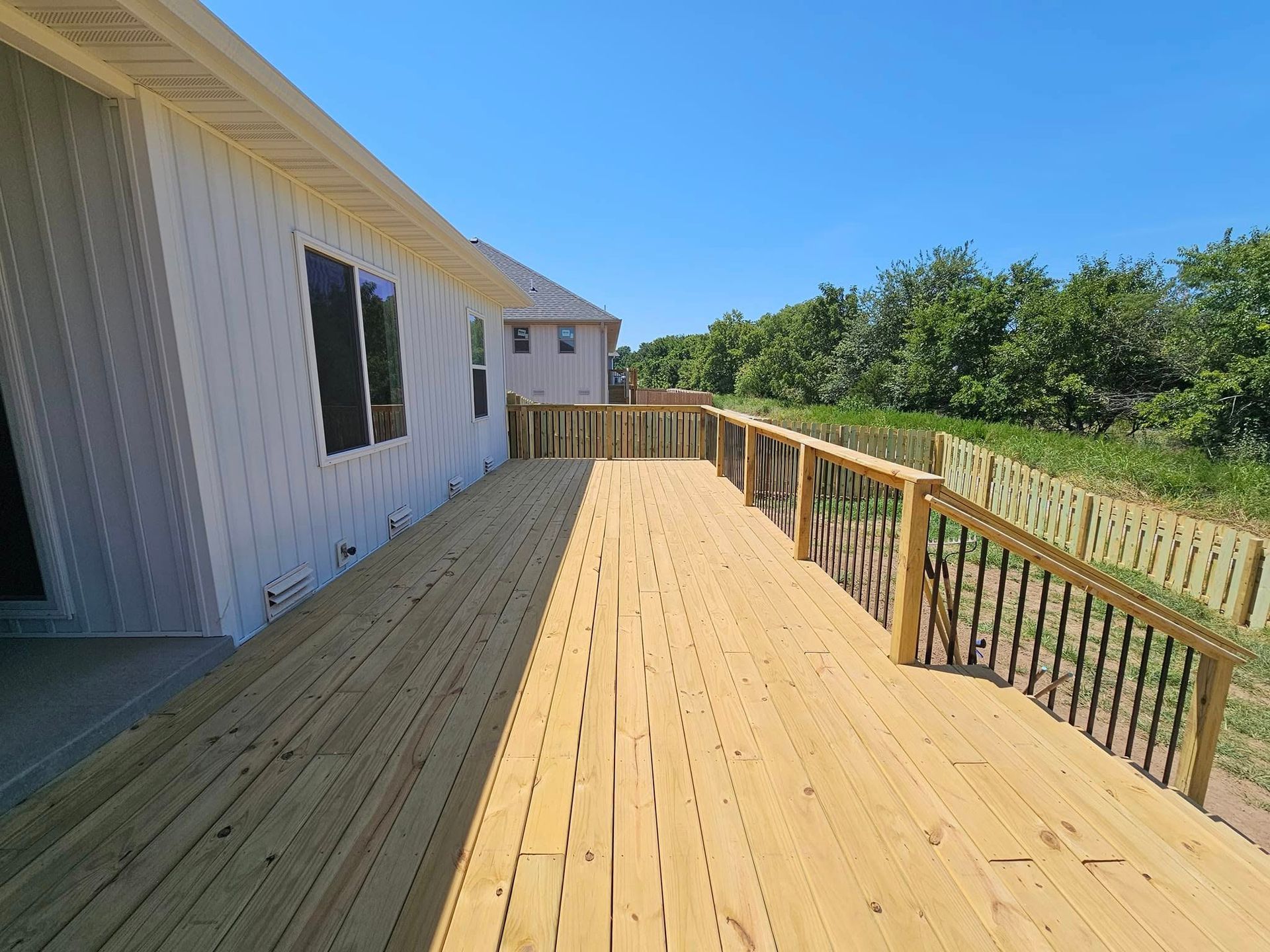 Wooden deck beside a white house, with railing and green trees under a bright blue sky.
