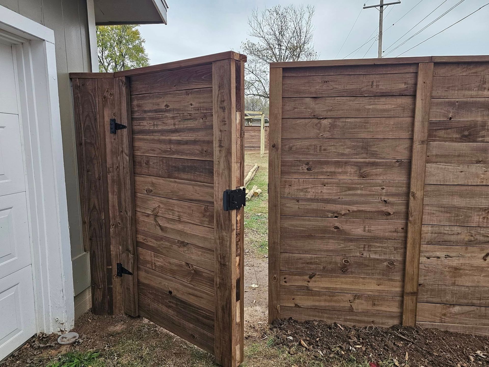 Wooden backyard gate with two hinged panels beside a white building, opening to a grassy yard.