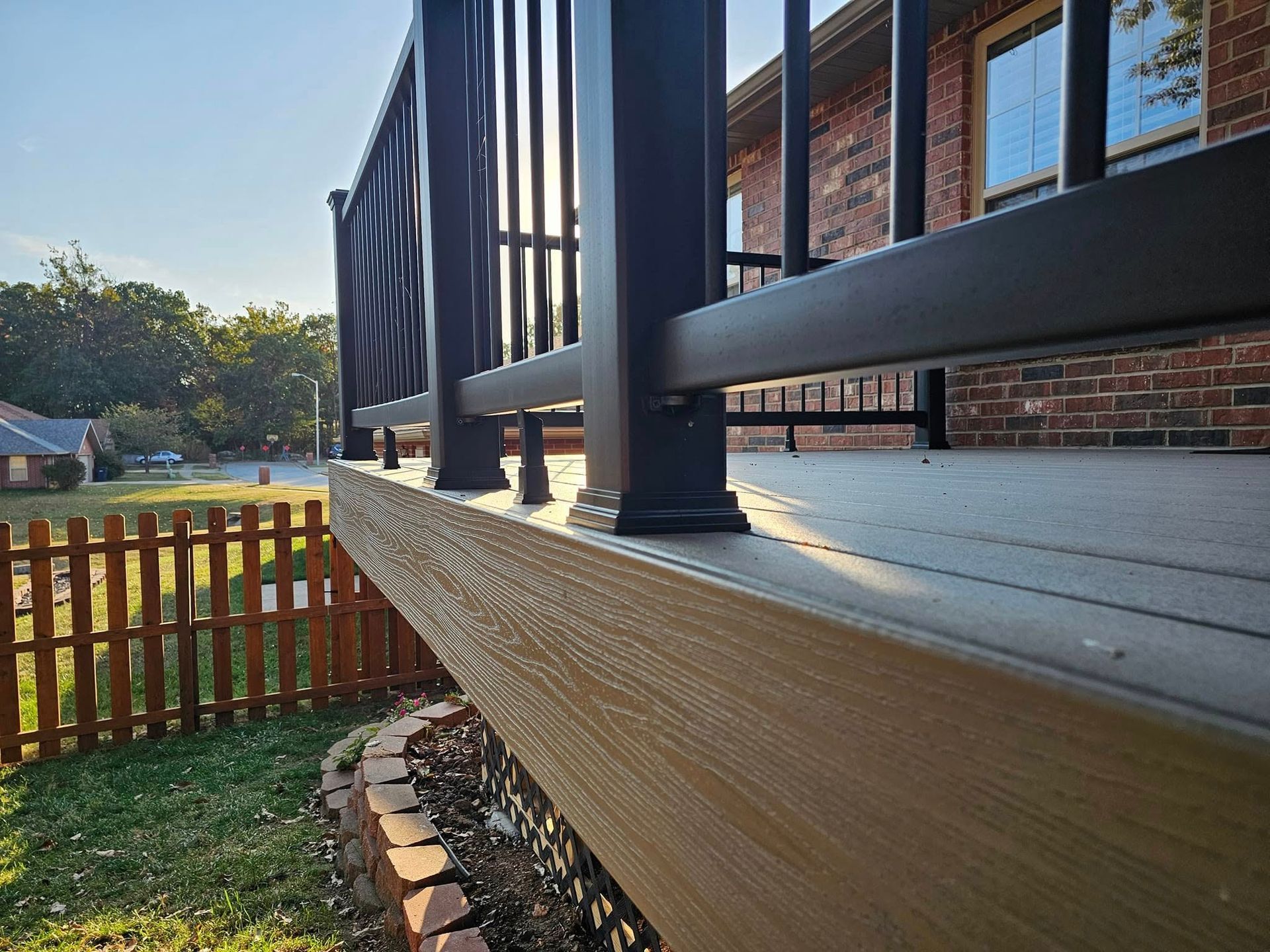 Side view of a wooden deck with black railing beside a brick house and fenced yard