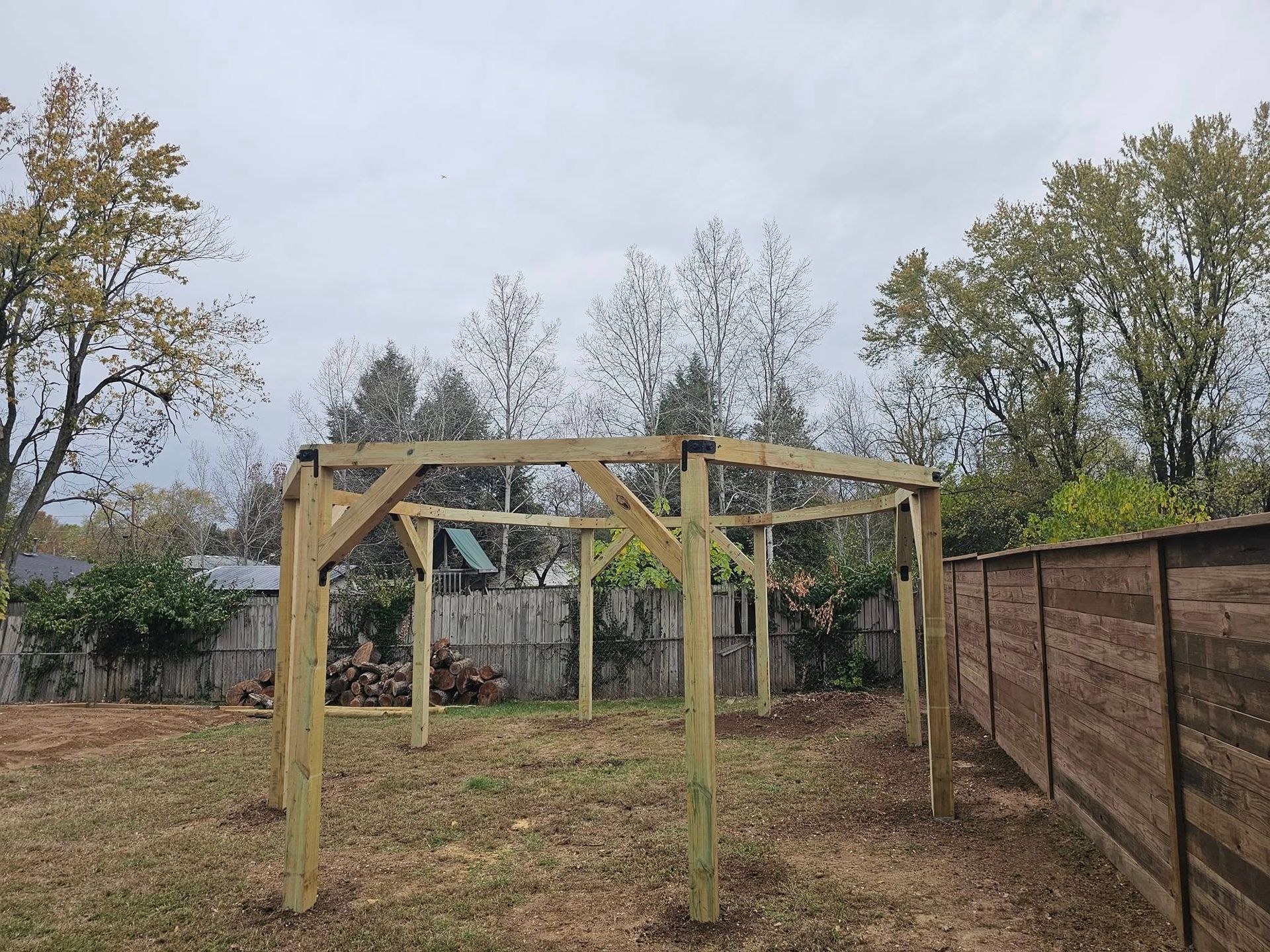 Wooden pergola frame in a backyard beside a fence under an overcast sky.