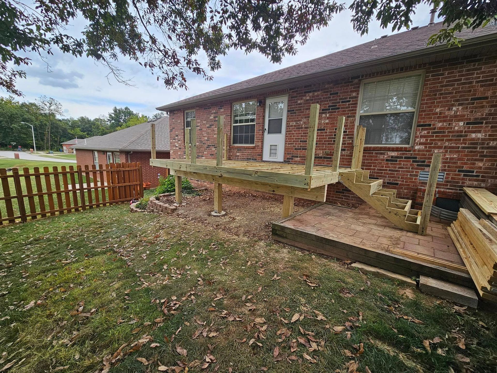 Back porch with wooden ramp and steps beside a brick house, fenced yard and fallen leaves.