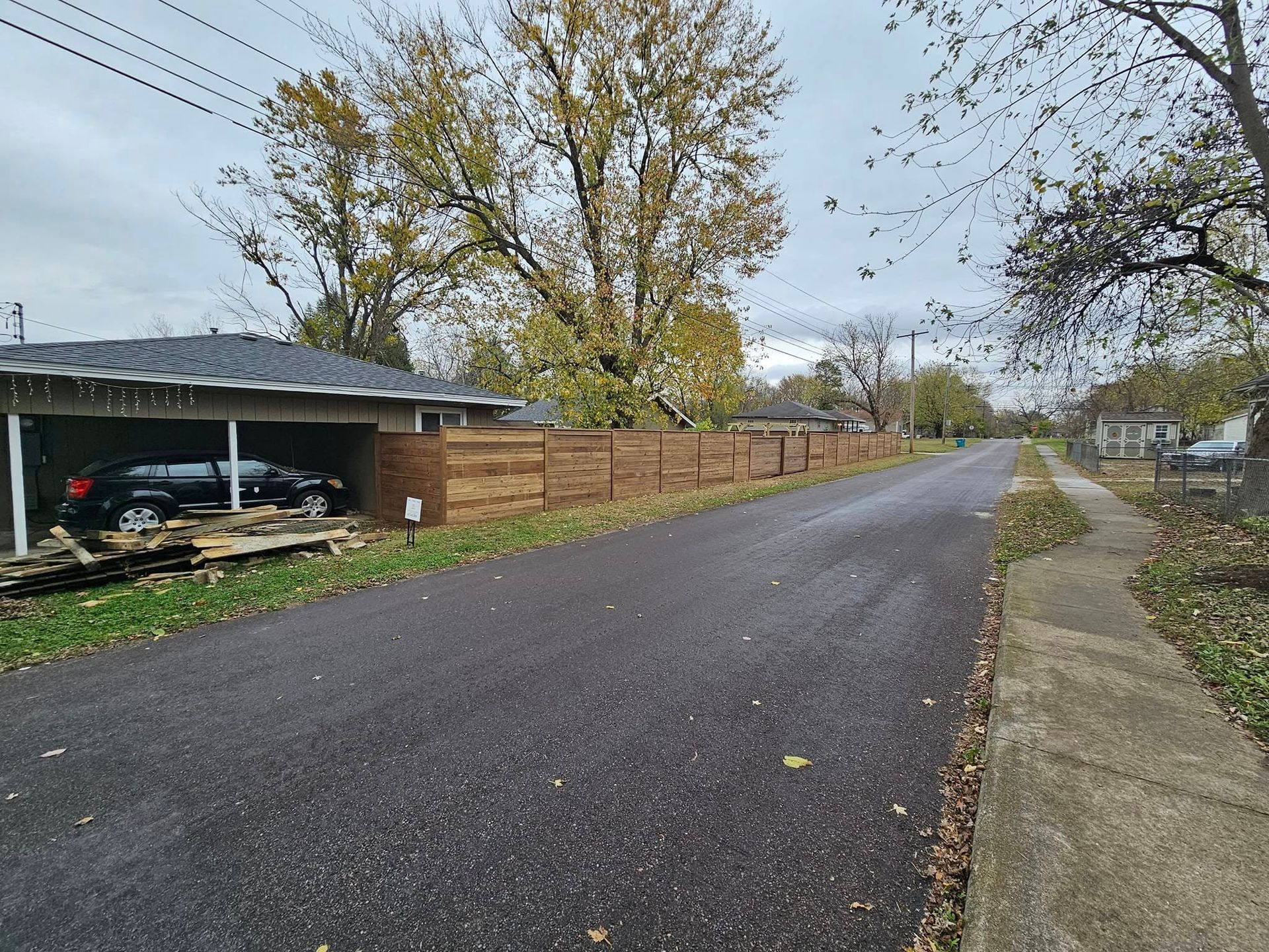 Quiet residential street with a parked car, trees, and a wooden fence on an overcast day