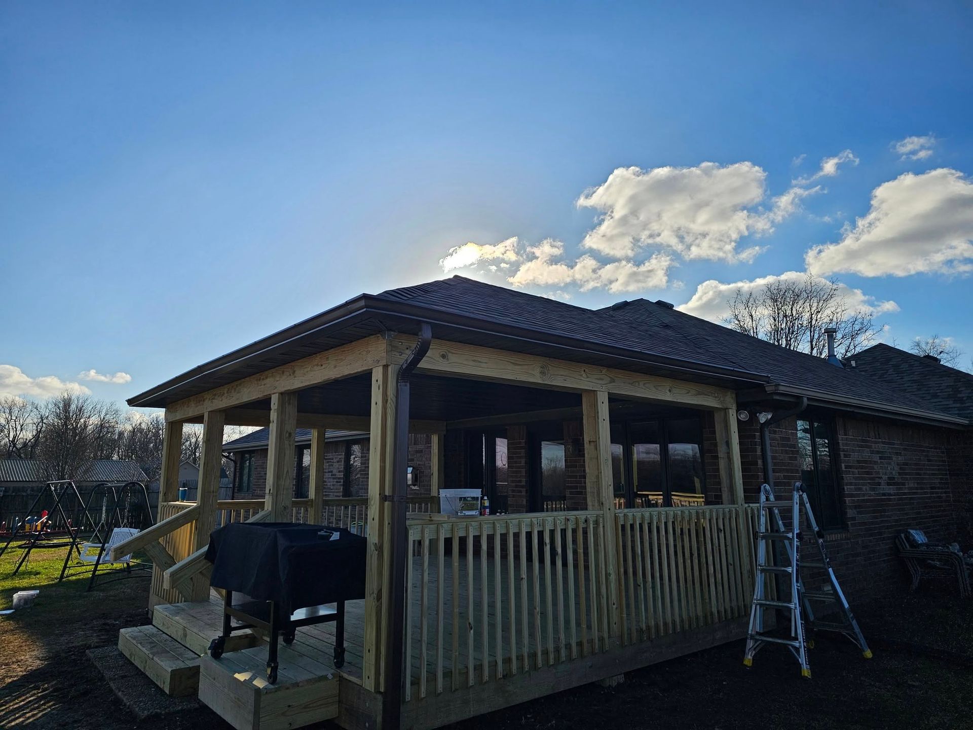 Backyard covered patio with wooden railing, grill, and ladder under a blue sky