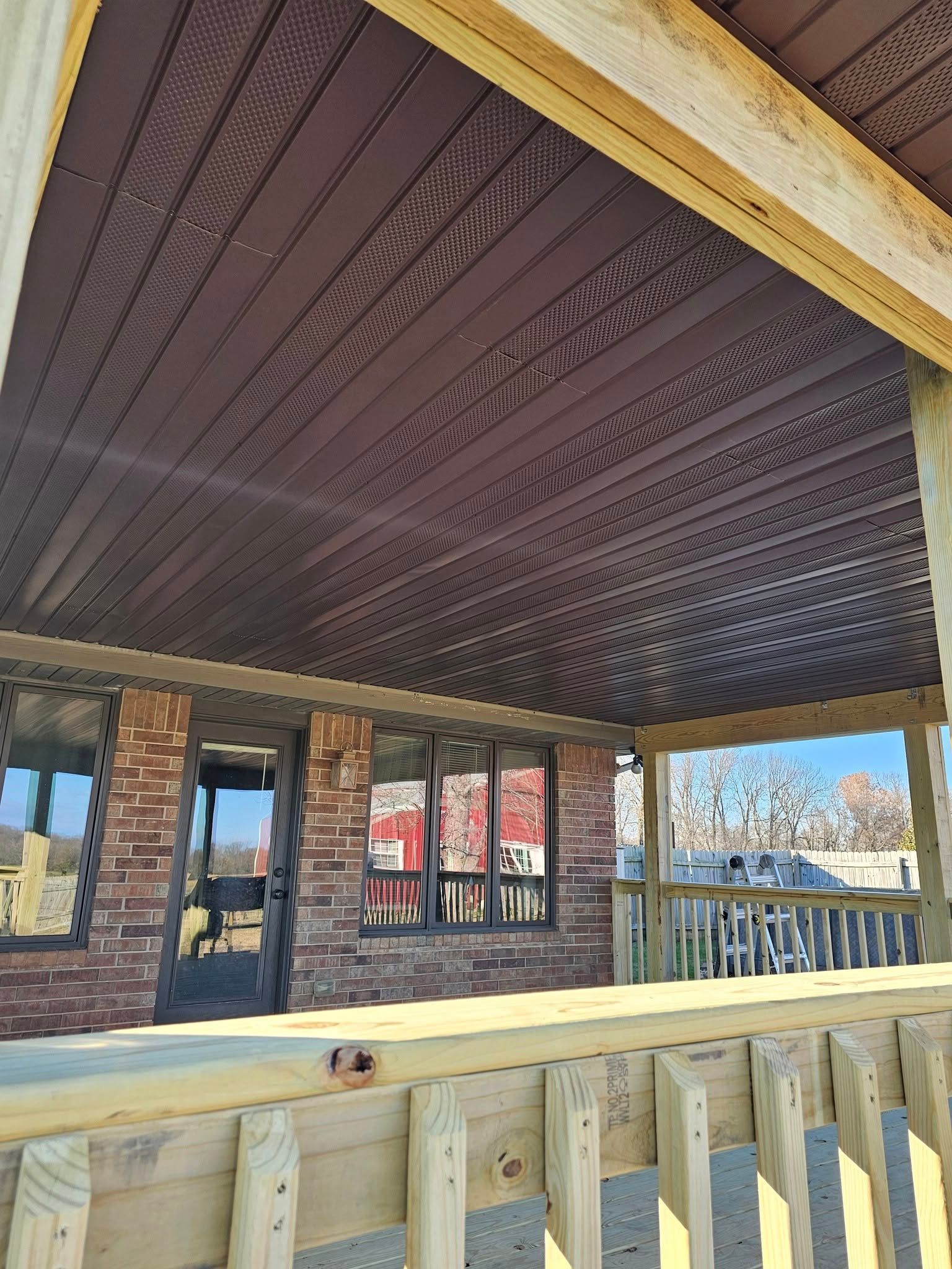 Covered patio with dark patterned ceiling, stone columns, and wooden railing overlooking a bright outdoor view