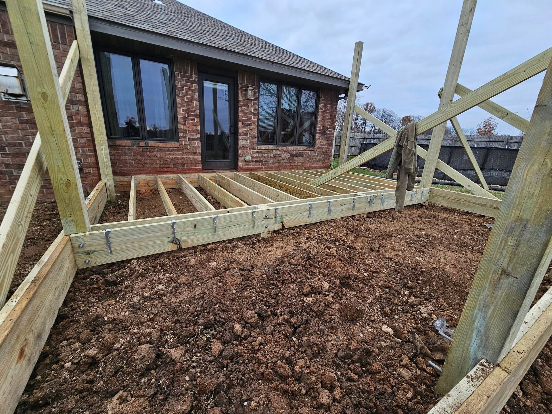 Wooden garden deck frame under construction beside a brick house, with soil in the foreground.