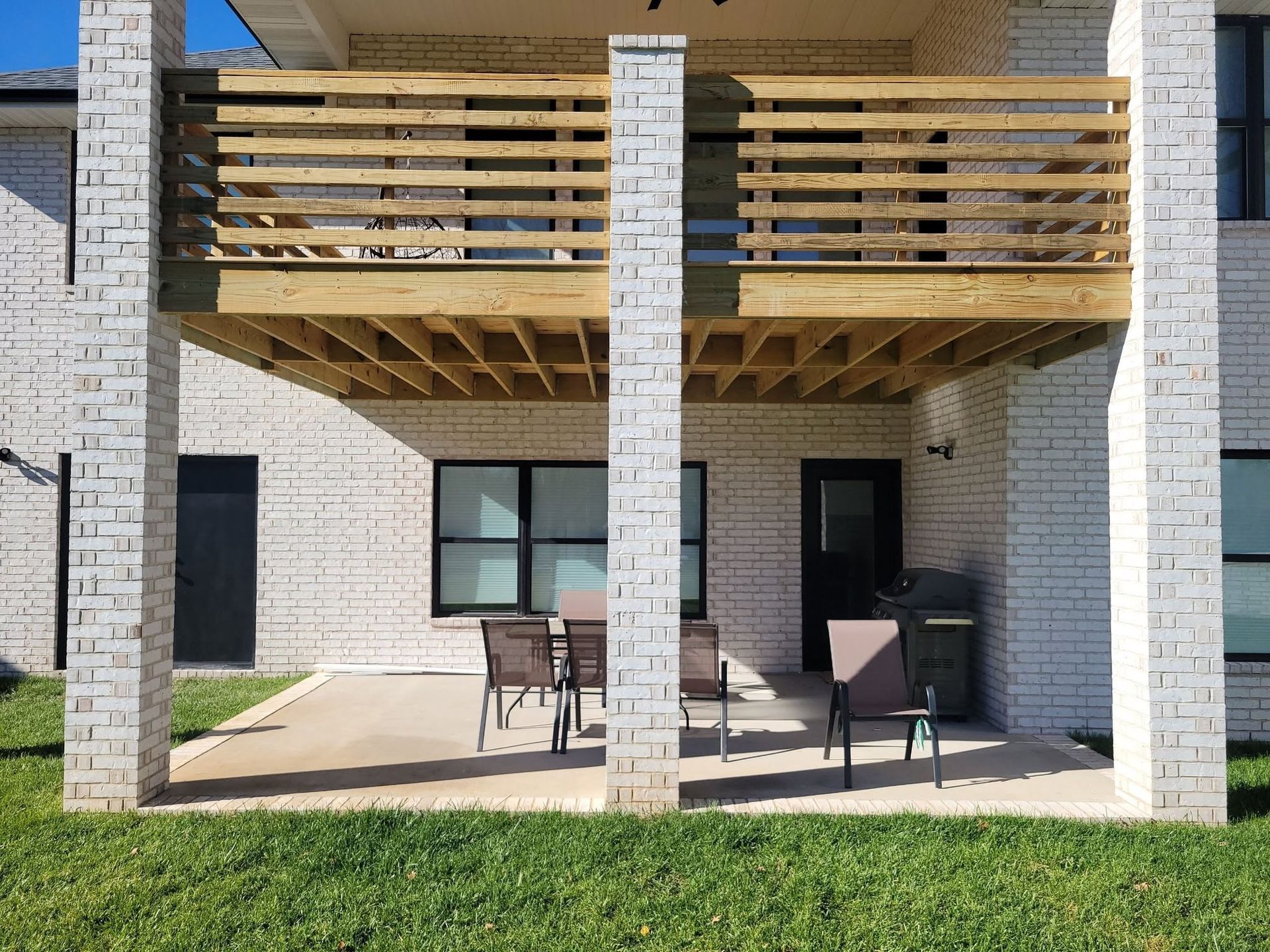Covered patio with brick columns, wooden balcony above, and outdoor chairs on a lawn.