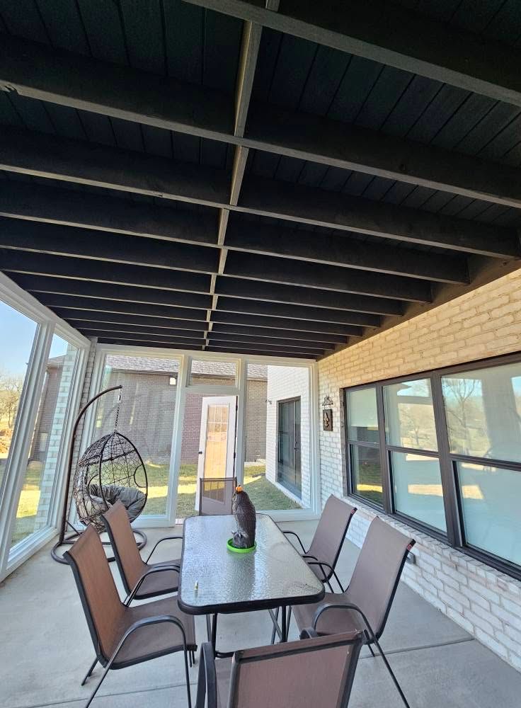 Covered patio with glass-top dining table, four chairs, and a standing fan beside windows.
