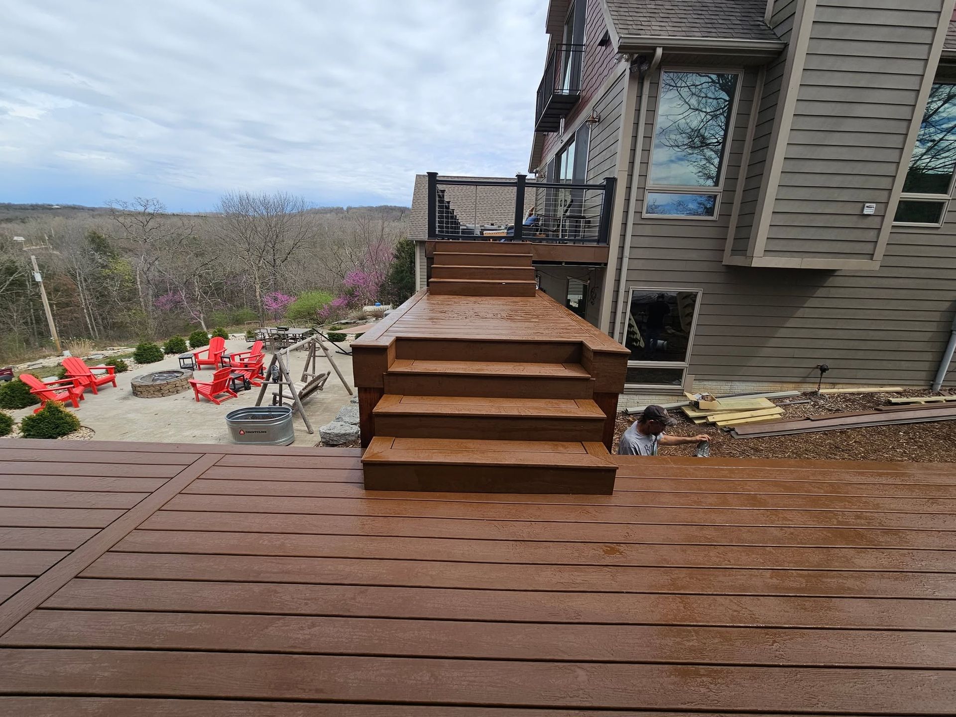 Wooden deck with steps leading to a house, overlooking a hillside yard with red chairs and planters.