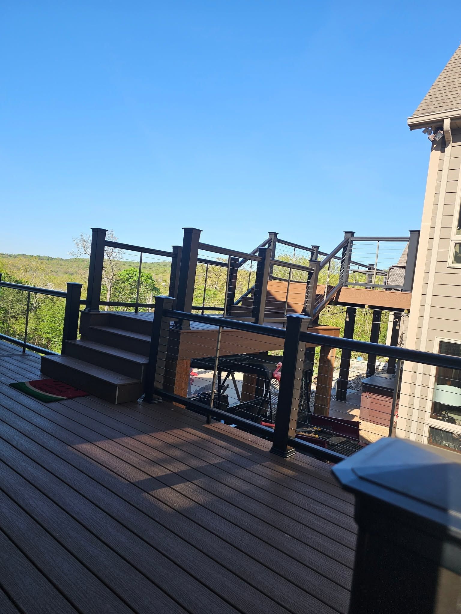 Wooden deck with black railing beside a brick house, overlooking a grassy landscape under a clear blue sky.