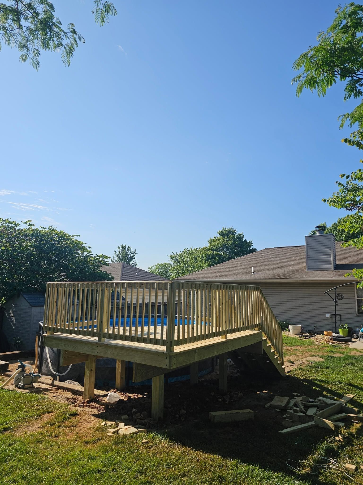 Wooden backyard deck under construction beside a house, with blue sky and trees overhead.