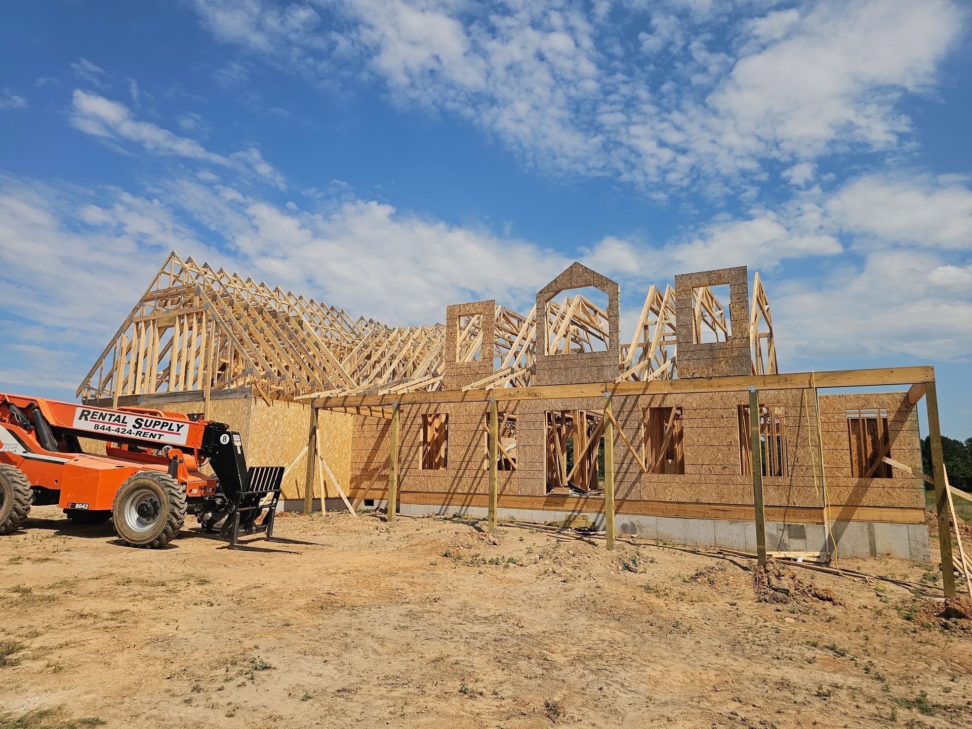 Partially built wooden house frame with a red tractor on a sandy lot under a blue sky