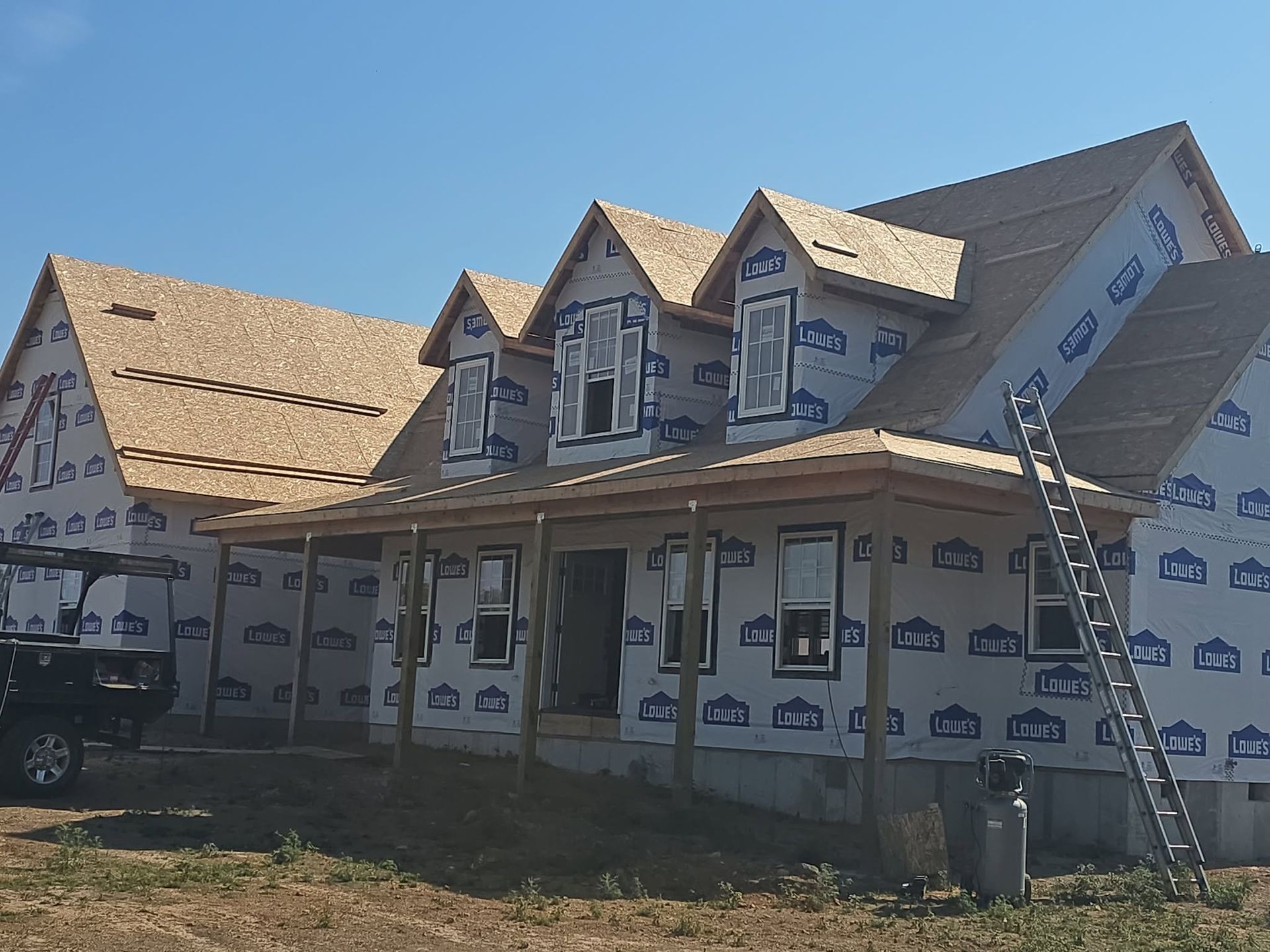 Partially built house with roofing, porch, ladder, and exposed house wrap under a clear sky