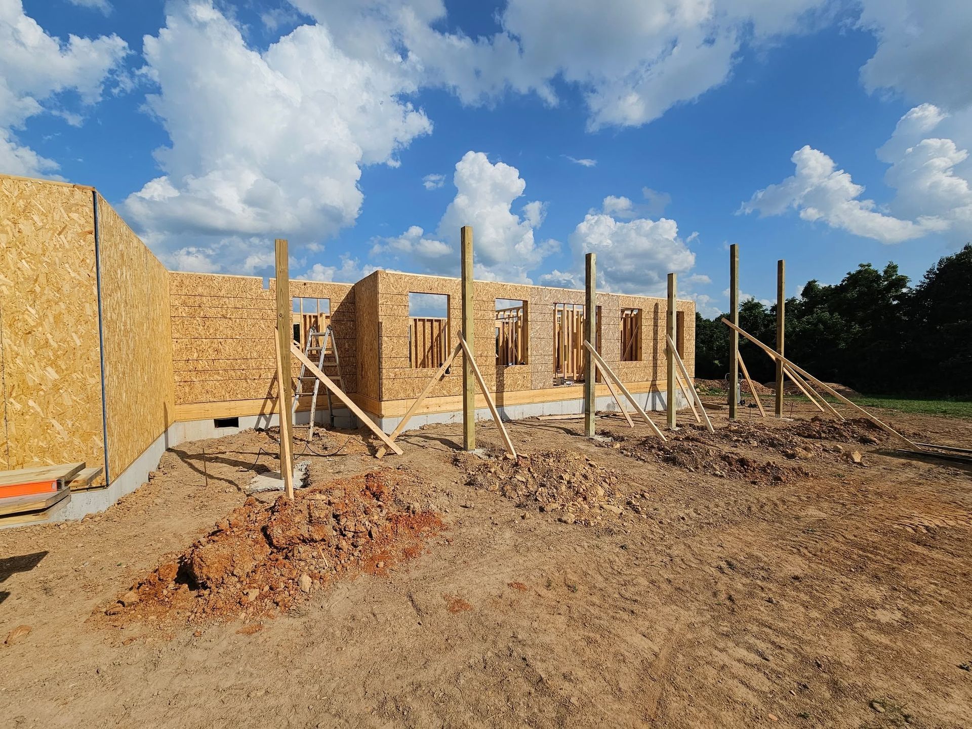 Wood-framed house under construction on a dirt lot beneath a partly cloudy blue sky