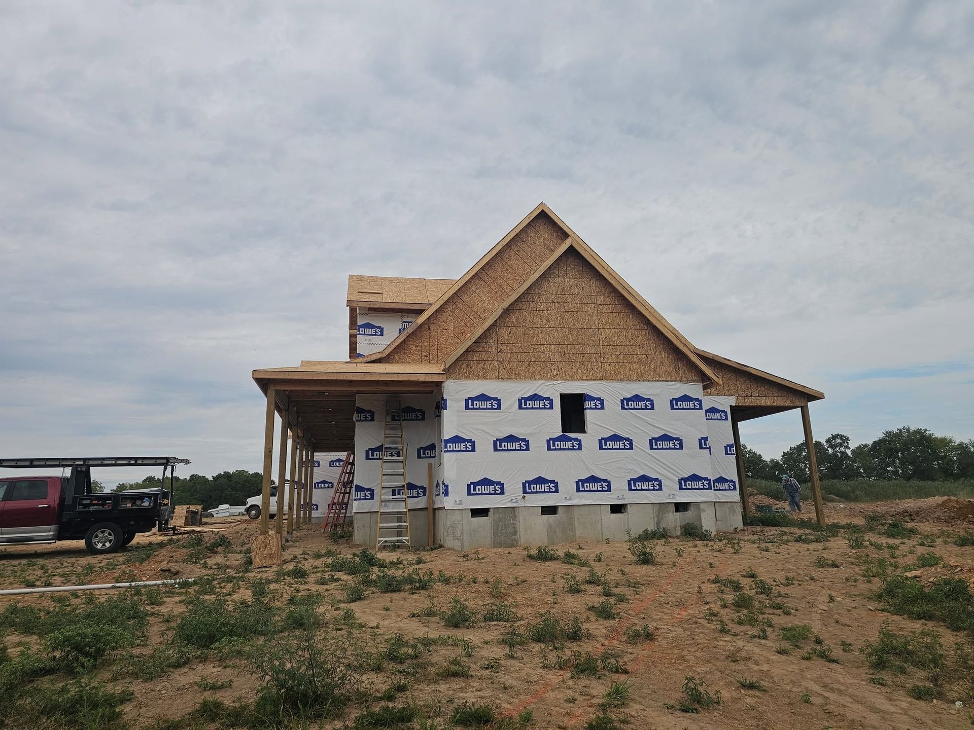 Partially built house with exposed roofing and white wrap, on a dirt lot under a cloudy sky