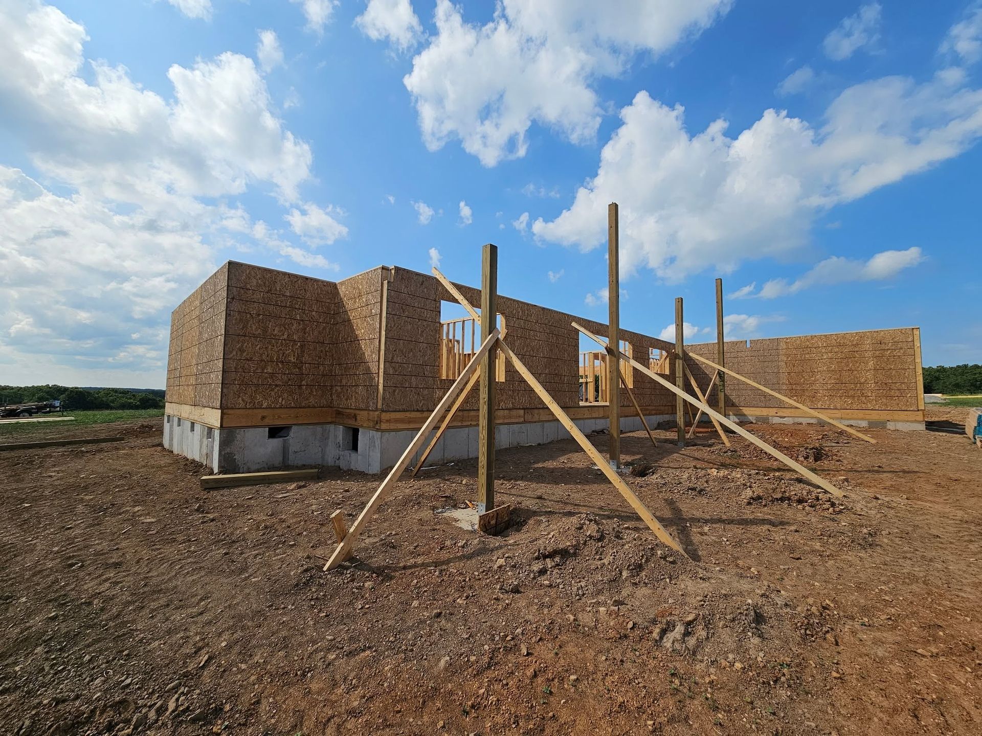 Wood-framed house under construction on a dirt lot beneath a blue, cloudy sky