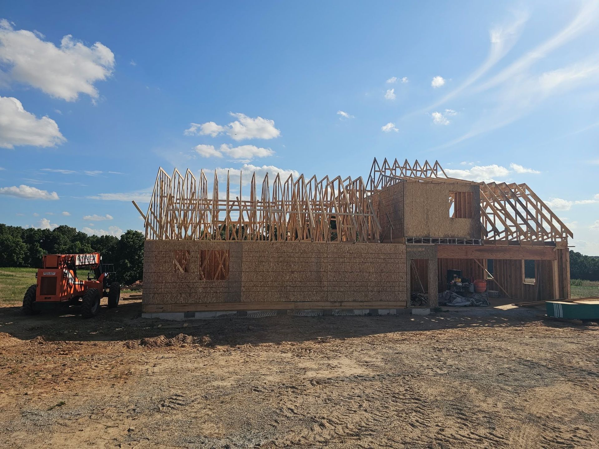 Wooden house under construction on a dirt lot, with roof trusses exposed and a red tractor nearby