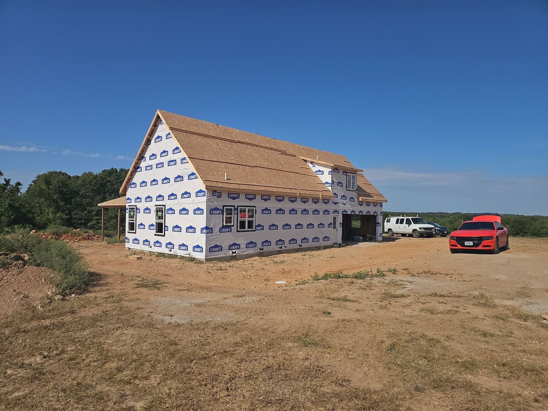 Partially built house wrapped in white housewrap, with a red car parked beside it on a dry lot.