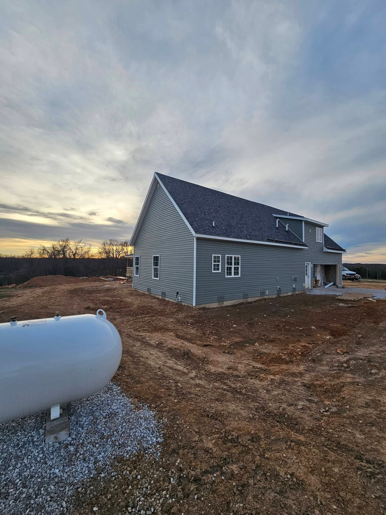 Small curved tiny house with dark roof on a gravel lot beside a white tank at sunset