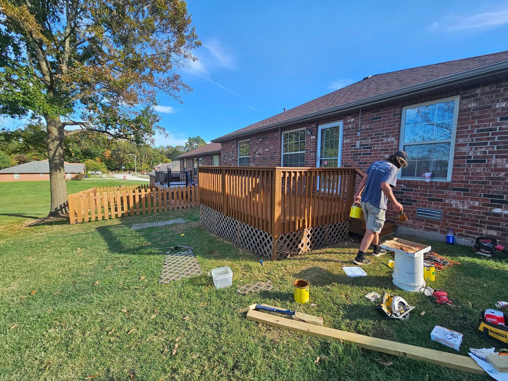 Person staining a wooden deck beside a brick house on a sunny day