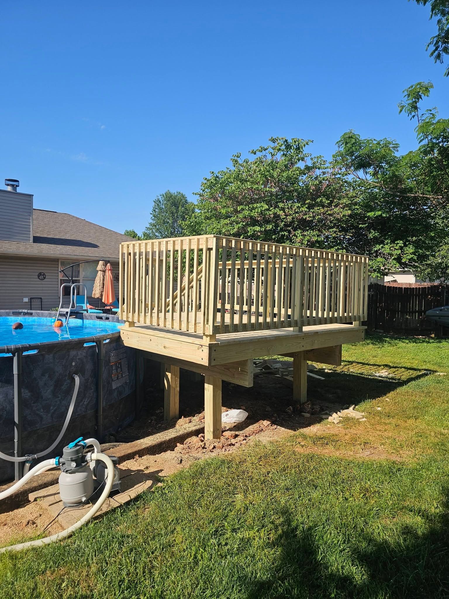 Small raised wooden deck under construction beside a backyard pool, with posts and railing in sunny grass.