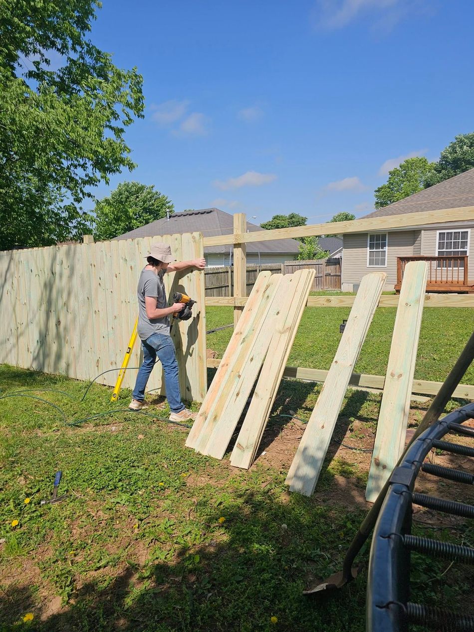 Person building a wooden fence in a sunny backyard, with boards leaning against posts