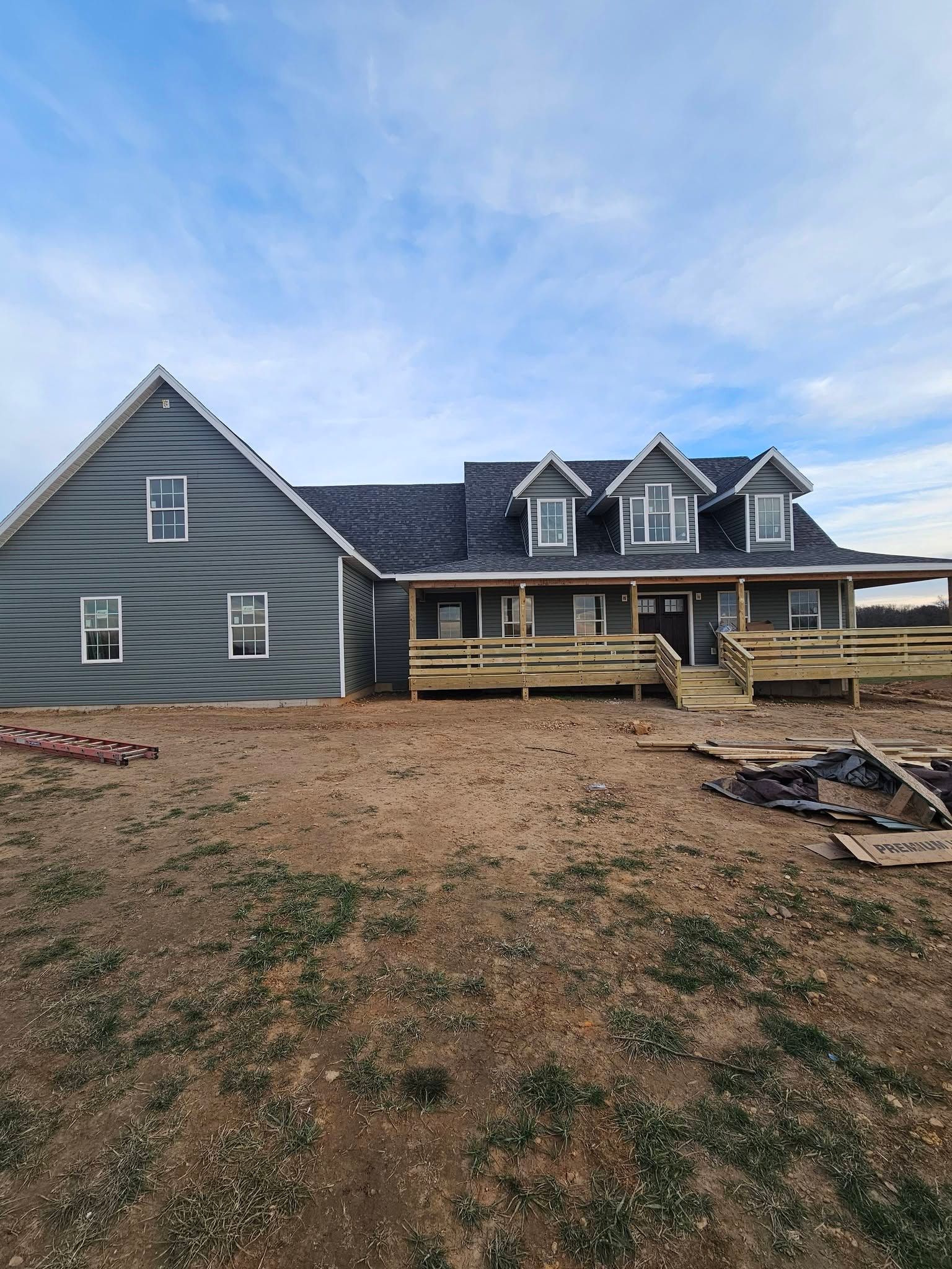 Large gray house under construction with unfinished porch and bare dirt yard beneath a cloudy sky
