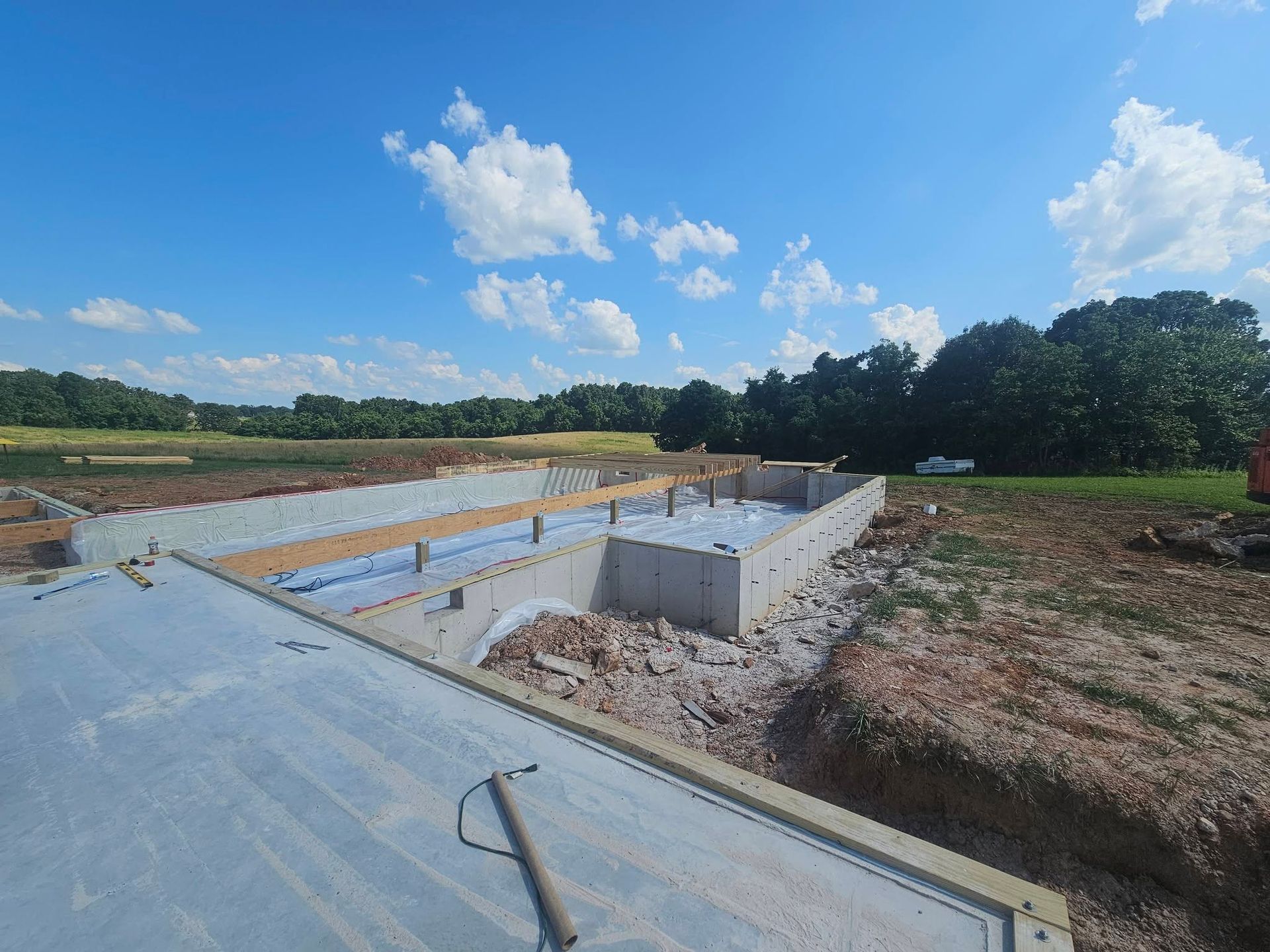 Construction site with concrete foundation walls and a wide blue sky over trees and dirt ground