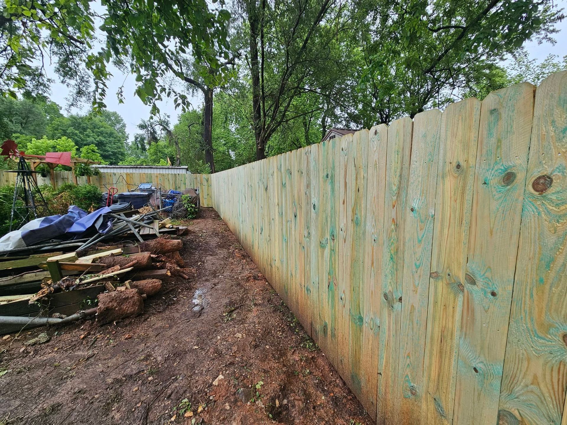 Wooden fence along a muddy yard with scattered debris and trees in the background