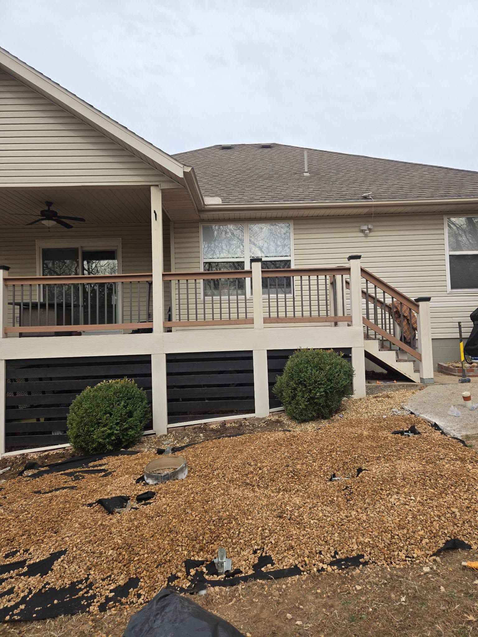 Single-story beige house with covered porch, black railings, and gravel landscaping under an overcast sky