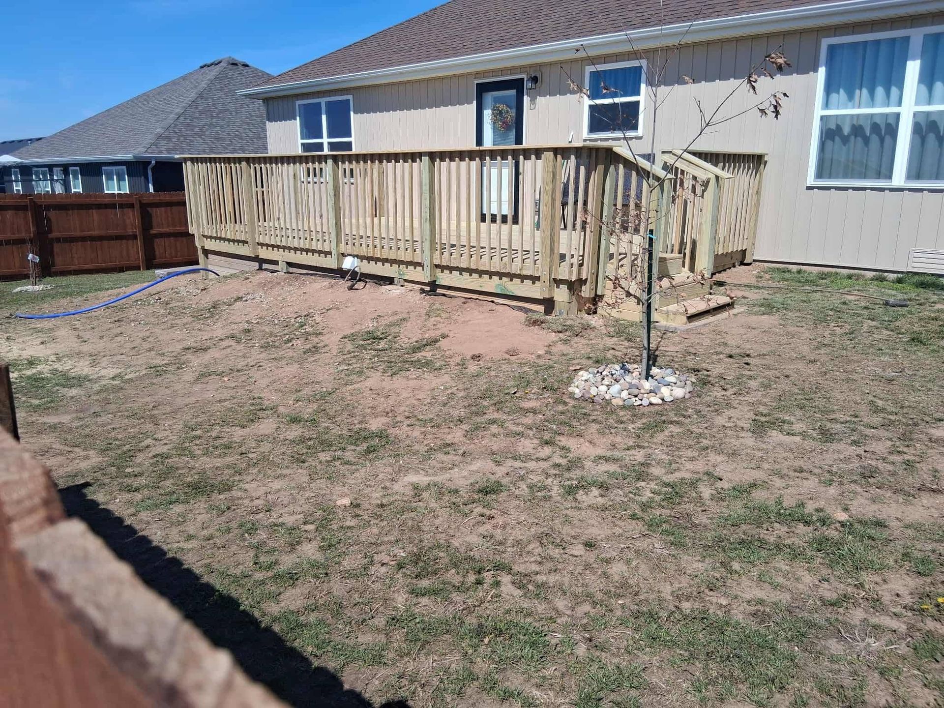 Backyard with wooden deck attached to a beige house, patchy grass, and a small tree in the yard