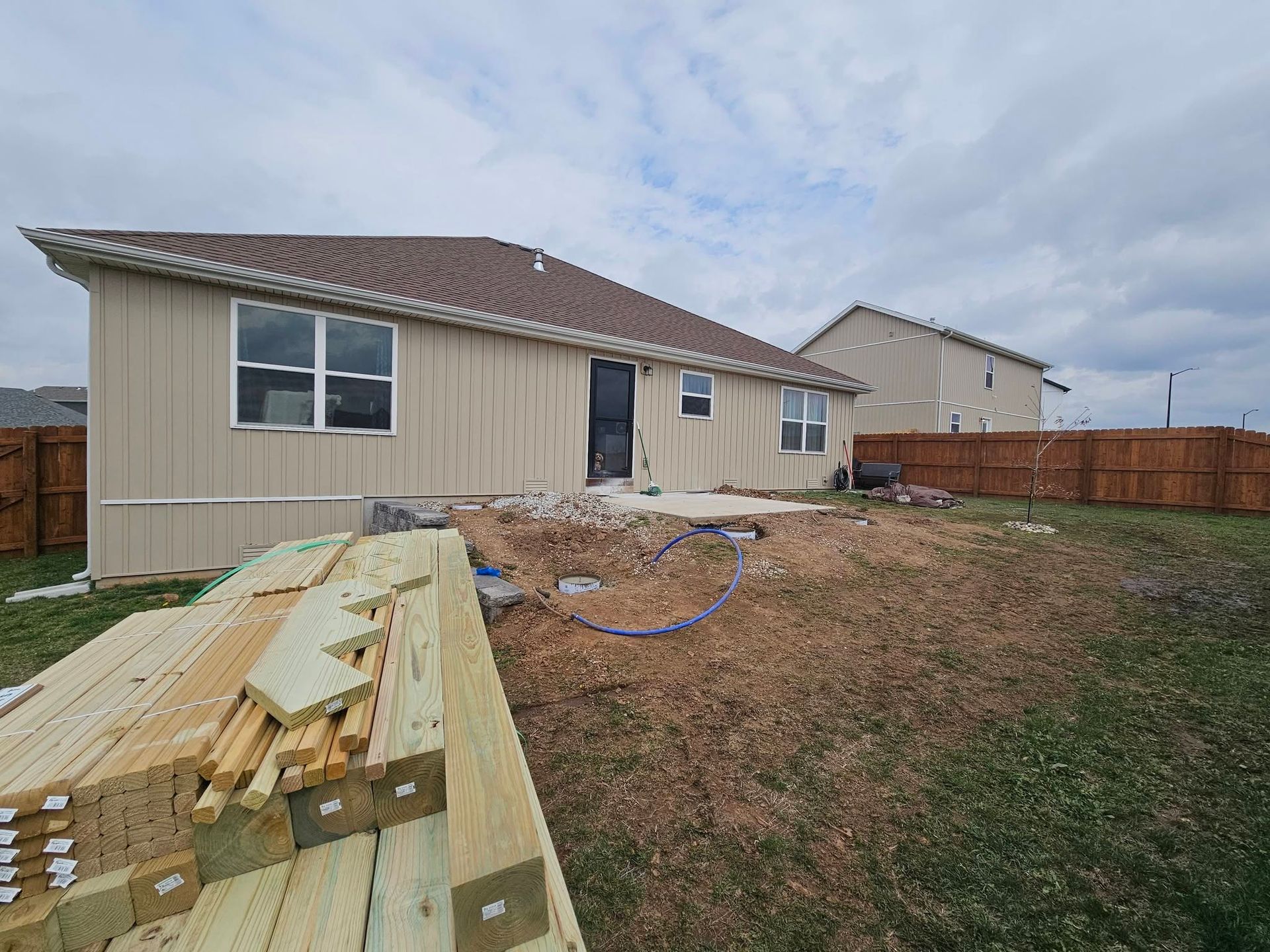 Backyard with house exterior, lumber piles, and fenced yard under cloudy sky