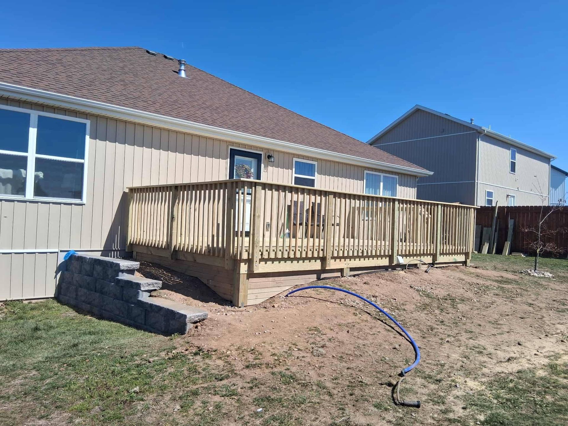 Backyard wooden deck attached to a tan house with stairs and a garden hose on the grass