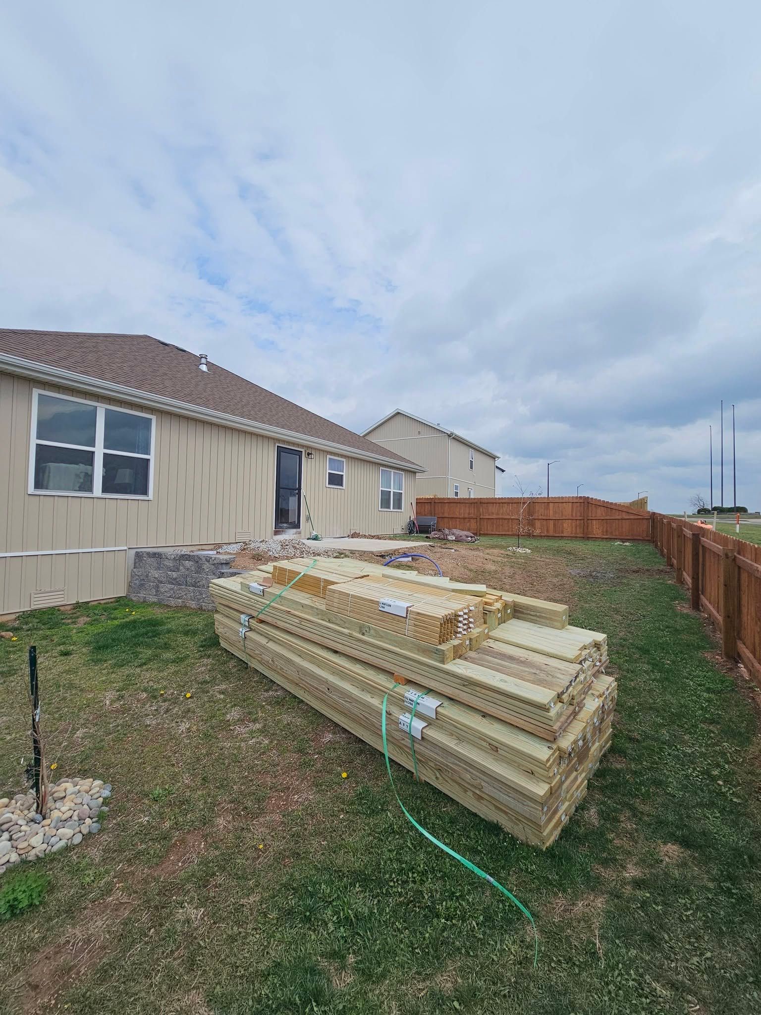 Stacked lumber beside a beige house in a suburban yard under a cloudy sky.
