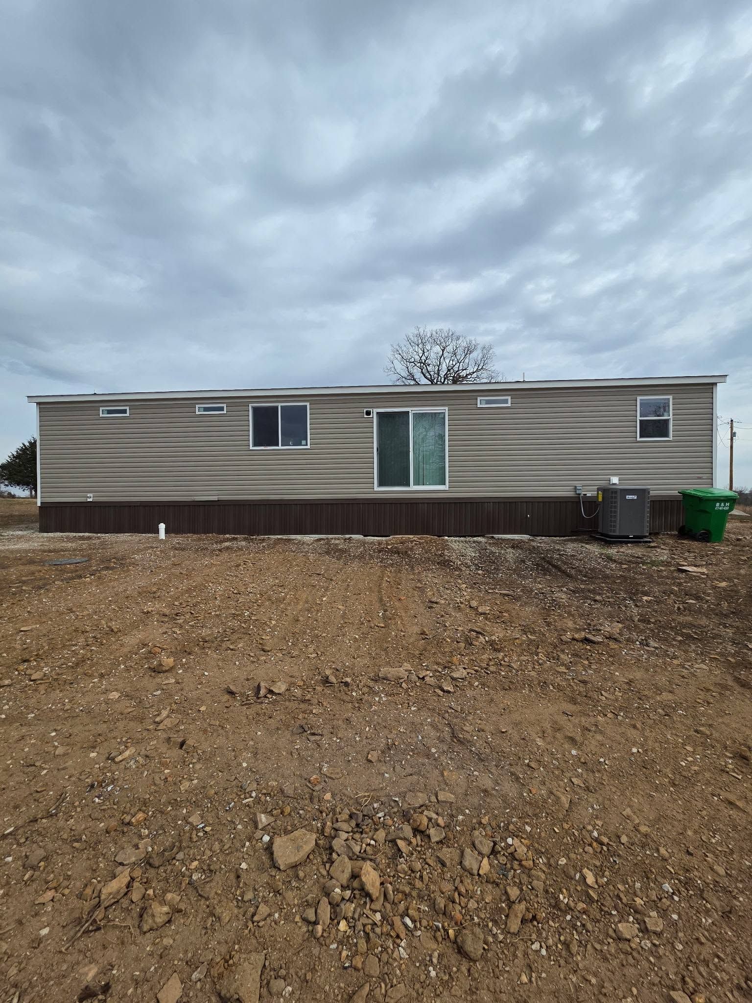 Mobile home on a dirt lot under a cloudy sky, with trash bins beside it.