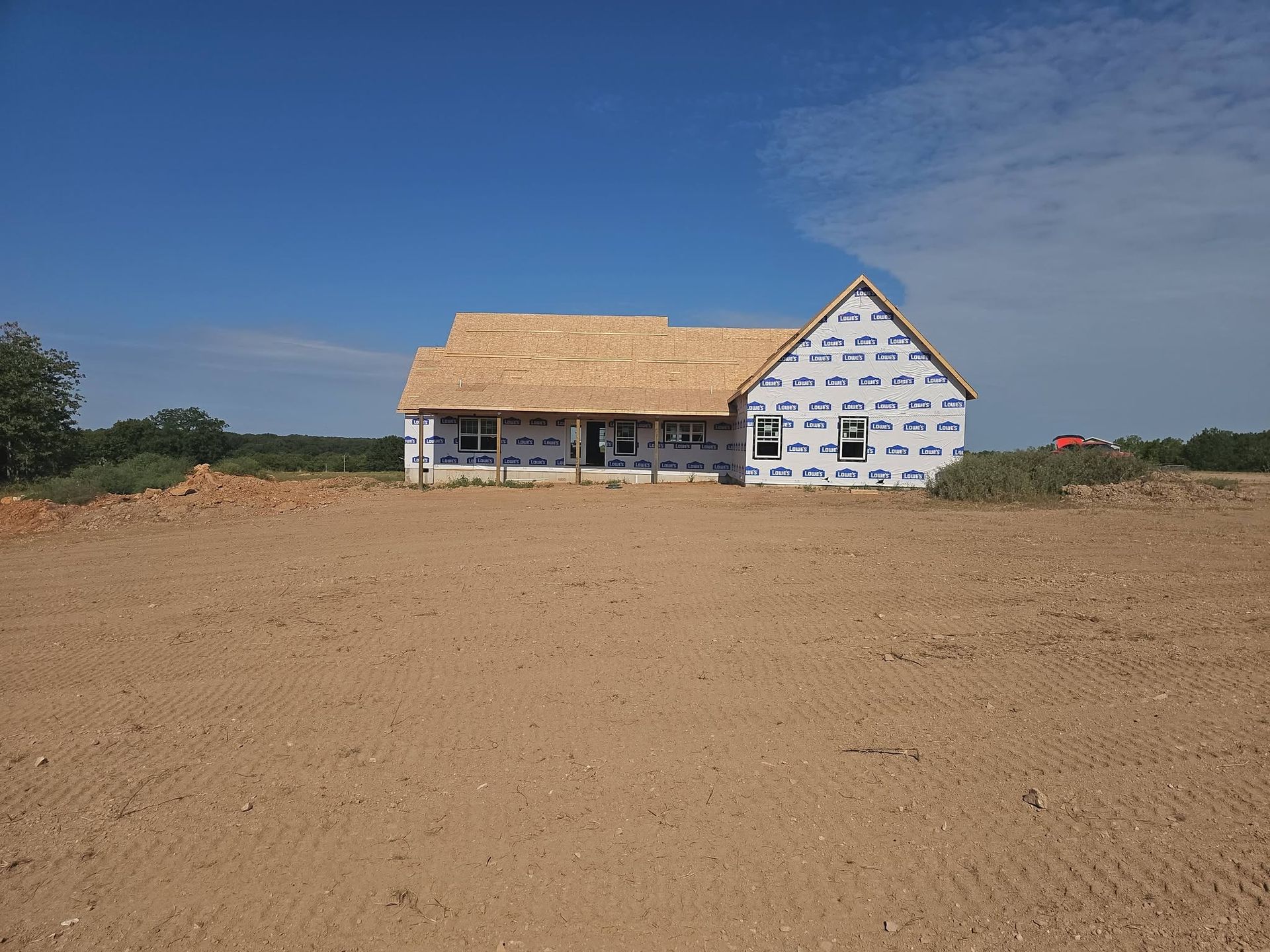 Partially built house with tan roof and white wrap on a barren dirt lot under a blue sky